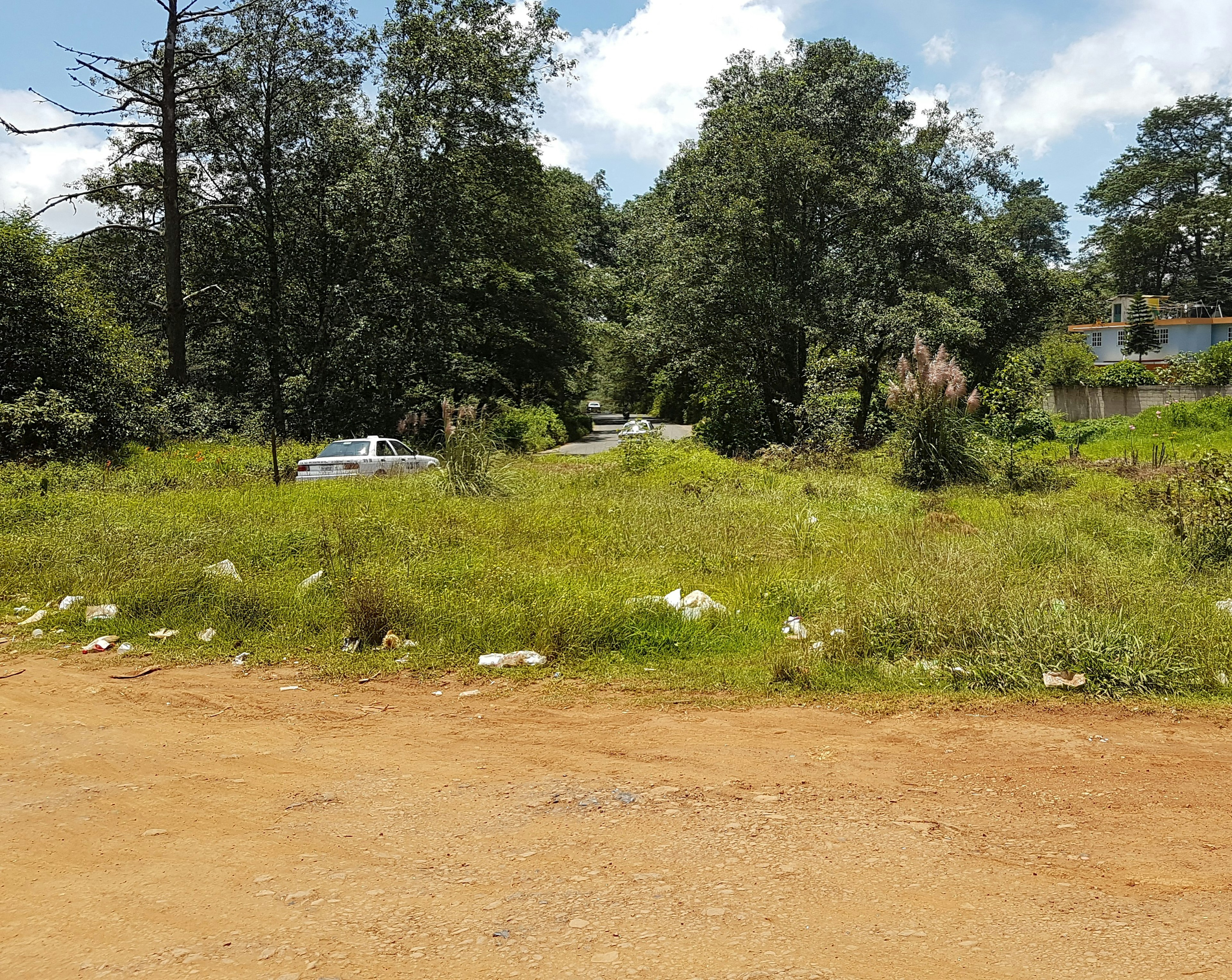 La imagen muestra un terreno sin desarrollar en una zona suburbana o rural. Se observa una carretera de tierra en primer plano, un área de césped con algo de basura, y un camino asfaltado rodeado de árboles al fondo. Hay un automóvil blanco visible, posiblemente un taxi. El entorno sugiere un área en desarrollo con potencial para futuros proyectos inmobiliarios o de urbanización.