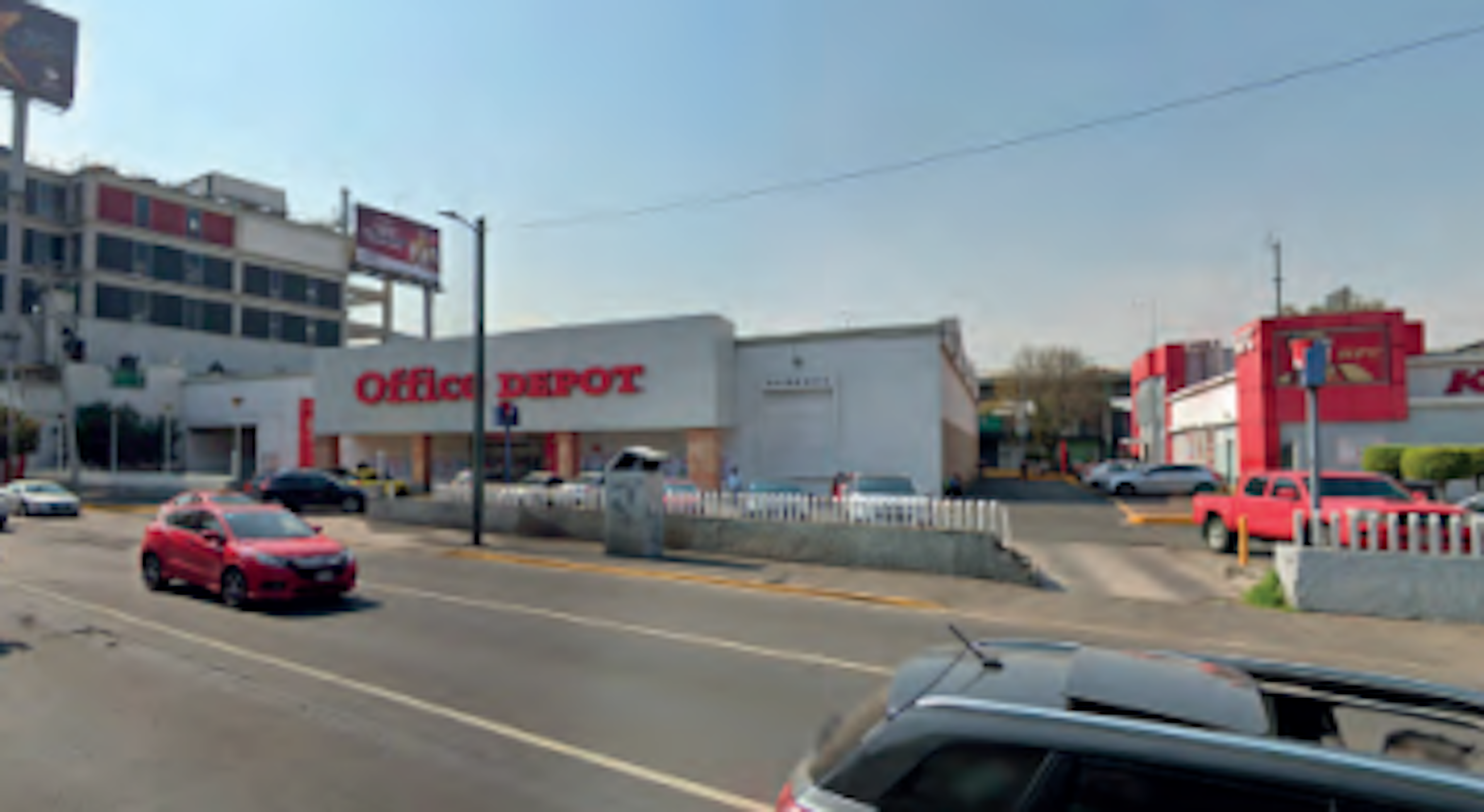 La imagen muestra una zona comercial con un Office Depot como edificio principal. El área parece ser un distrito de negocios suburbano o de las afueras de una ciudad. Hay varios vehículos estacionados y circulando por la calle. El entorno incluye otros edificios comerciales y señalización publicitaria. La escena sugiere una zona de alto tráfico comercial típica de áreas suburbanas en desarrollo.