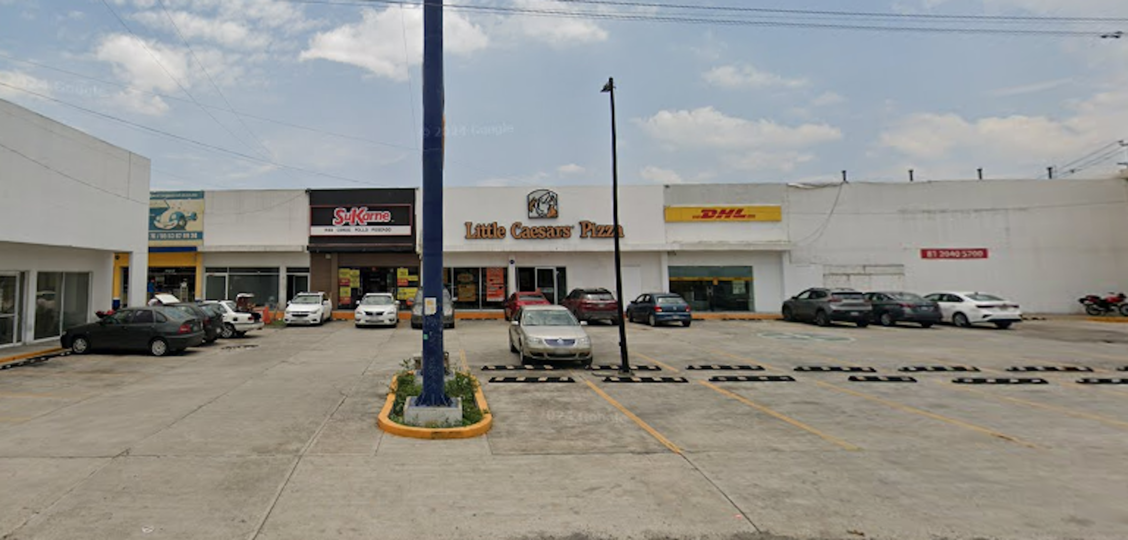 The image shows a small commercial strip mall with various businesses including Little Caesars Pizza, DHL, and other stores. The building is white with a parking lot in front filled with several cars. It appears to be a typical suburban or small town commercial property with retail and service establishments.