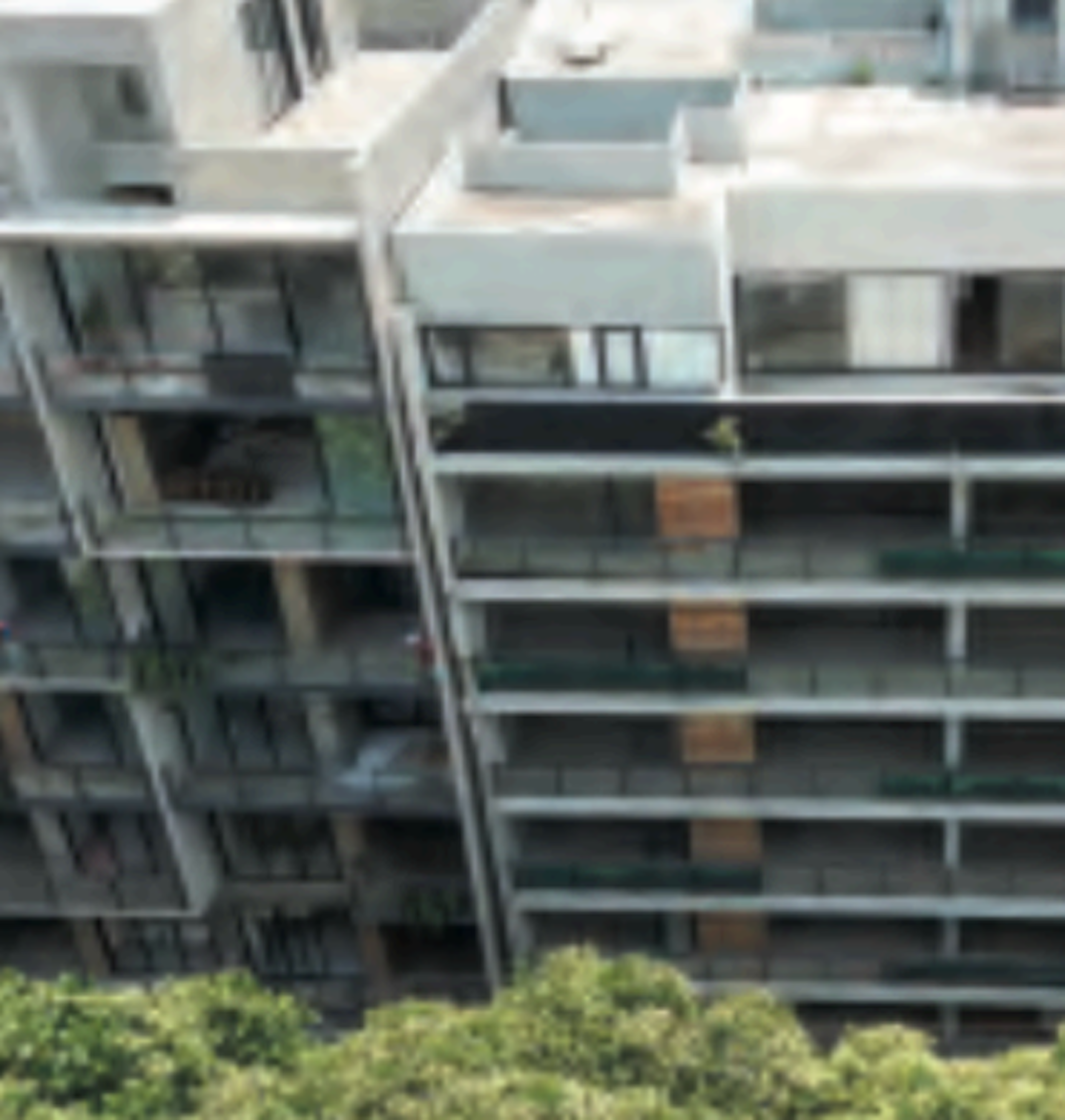 La imagen muestra un edificio de apartamentos moderno de varios pisos con balcones acristalados. La fachada es predominantemente blanca con detalles en gris. Se aprecian múltiples niveles y una arquitectura contemporánea. En la parte inferior de la imagen se ven algunas áreas verdes, sugiriendo un entorno residencial. La vista parece ser desde un punto elevado, posiblemente desde otro edificio cercano.