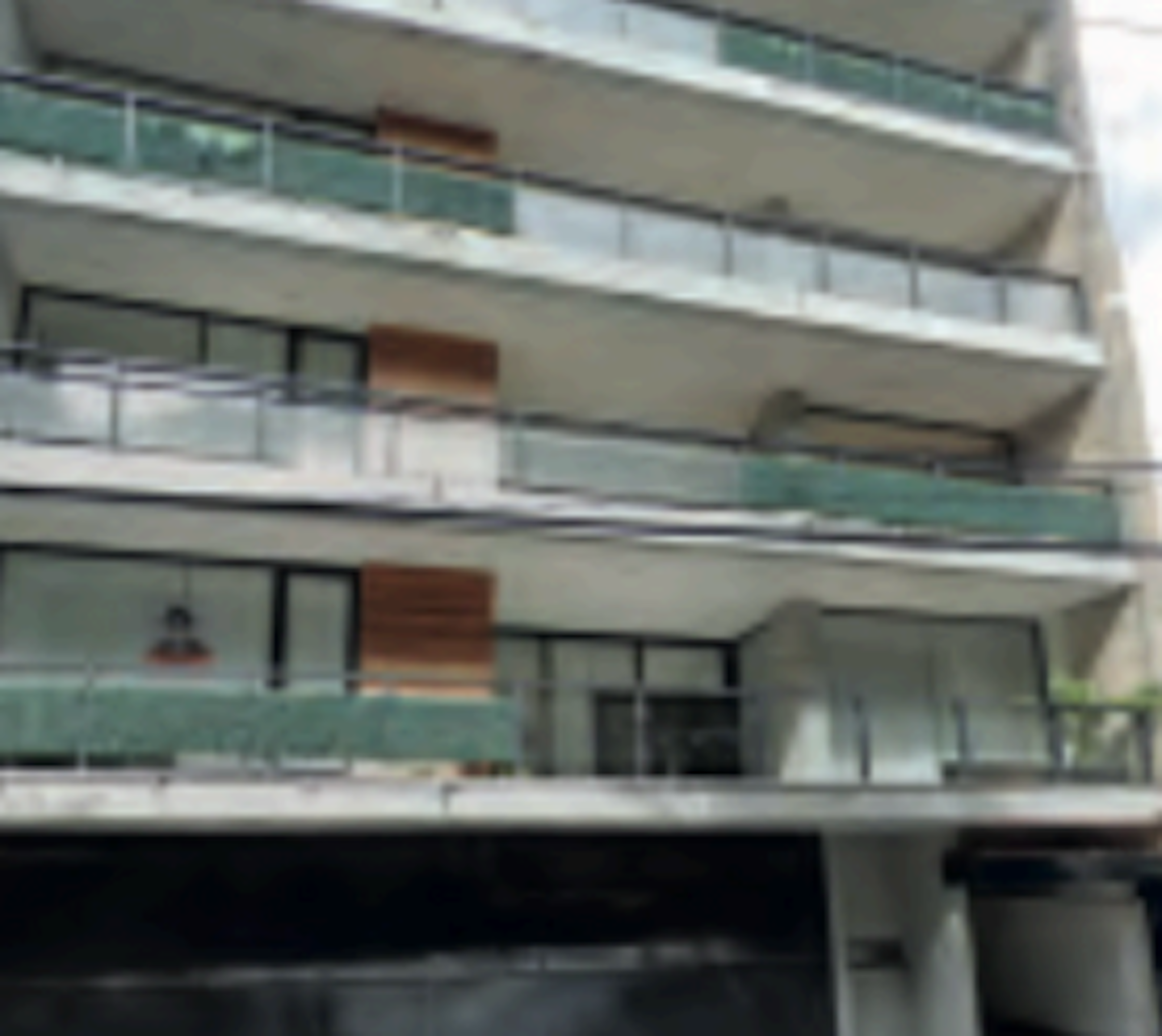 La imagen muestra la fachada de un edificio de apartamentos moderno con varios pisos. Se observan balcones amplios con barandillas de vidrio y metal. La estructura tiene un diseño contemporáneo con líneas limpias y una combinación de materiales como concreto, vidrio y elementos en madera. Los balcones parecen espaciosos y ofrecen vistas al exterior. En la planta baja se aprecia una entrada con puertas de vidrio.