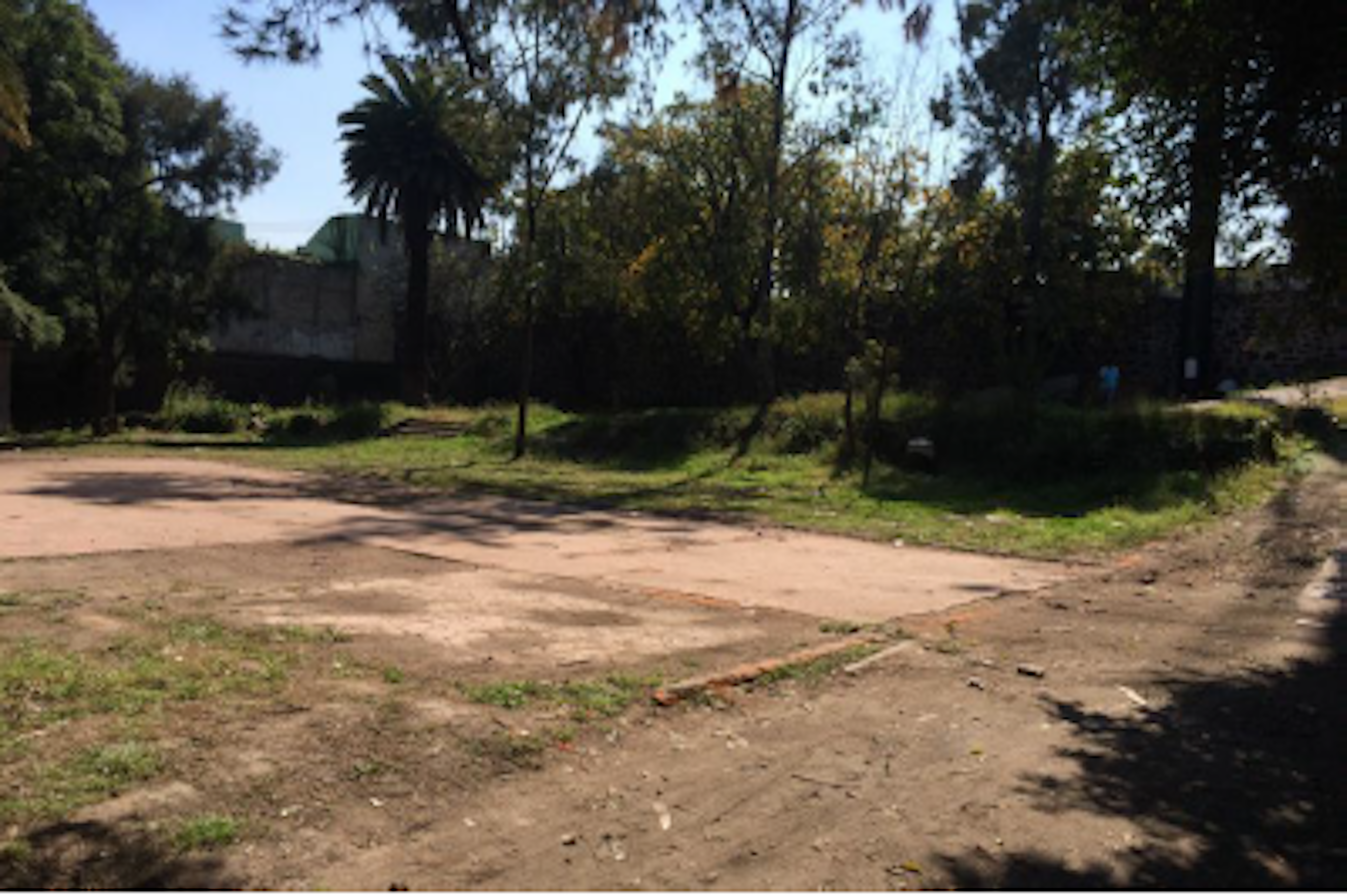 La imagen muestra un terreno baldío en una zona urbana. Se observa un área de tierra con algunas secciones pavimentadas, rodeada de vegetación y árboles, incluyendo una palmera. Al fondo se ve una pared o muro que delimita el terreno. El lugar parece descuidado y podría ser un espacio potencial para desarrollo inmobiliario o un proyecto de renovación urbana.