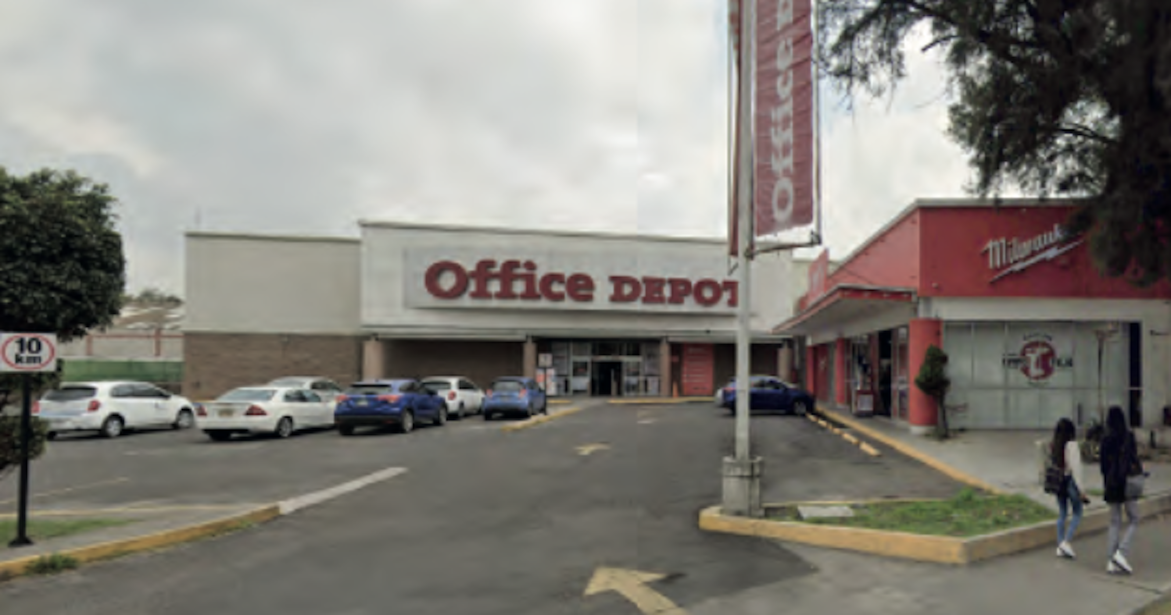 La imagen muestra un centro comercial suburbano con un Office Depot como tienda ancla. El estacionamiento está parcialmente lleno, indicando actividad comercial. El edificio es de un solo piso con fachada blanca y roja. Hay otras tiendas adyacentes, incluyendo una que parece ser Milwaukee. La escena sugiere una zona comercial típica en un área residencial o suburbana.