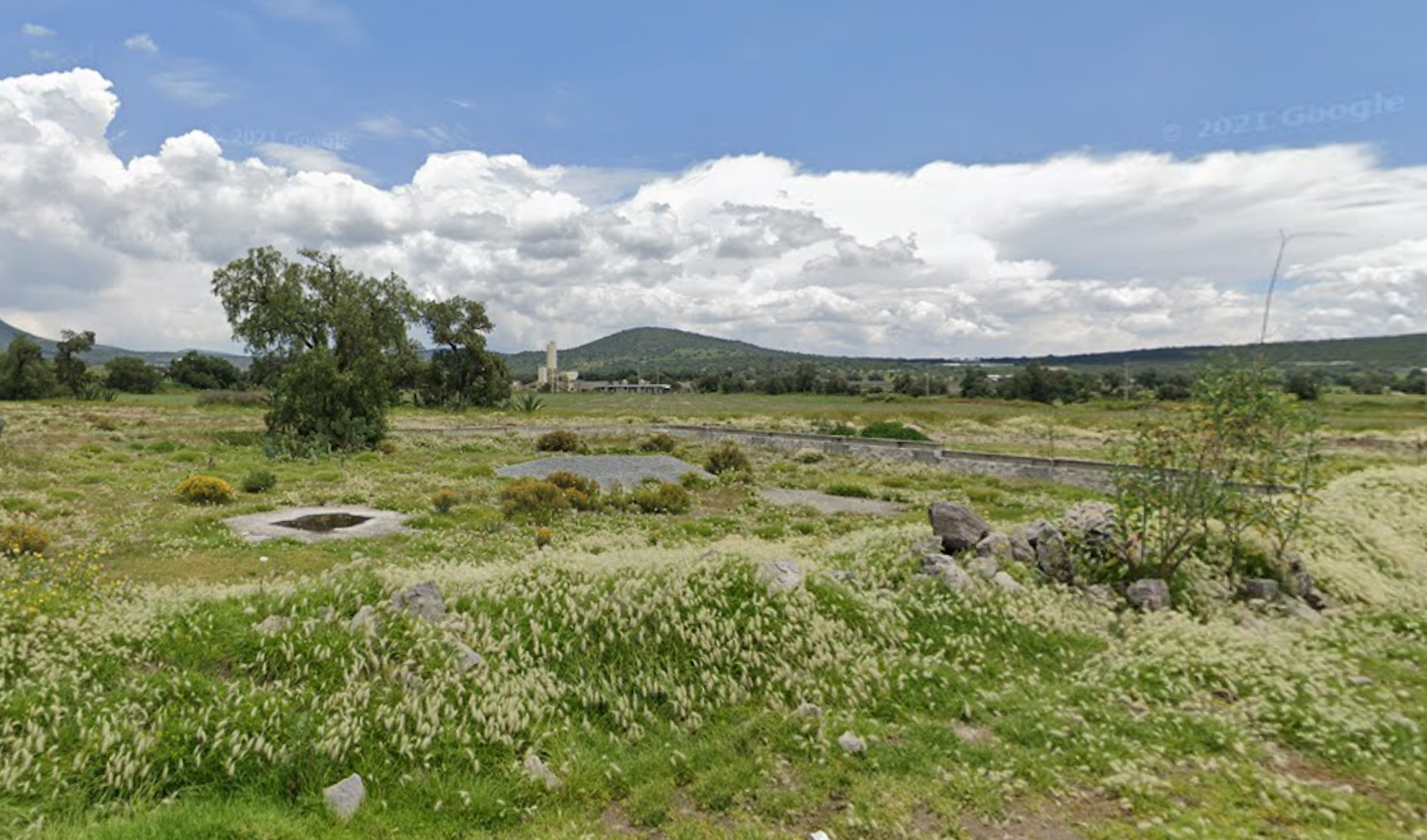 La imagen muestra un terreno rural amplio y sin desarrollar con vegetación silvestre. En el fondo se aprecian colinas y montañas bajas. Hay algunos árboles dispersos y lo que parece ser una estructura lejana, posiblemente un silo o torre. El cielo está parcialmente nublado. El terreno parece ser adecuado para desarrollo agrícola o potencialmente para proyectos inmobiliarios rurales, dependiendo de las regulaciones locales.