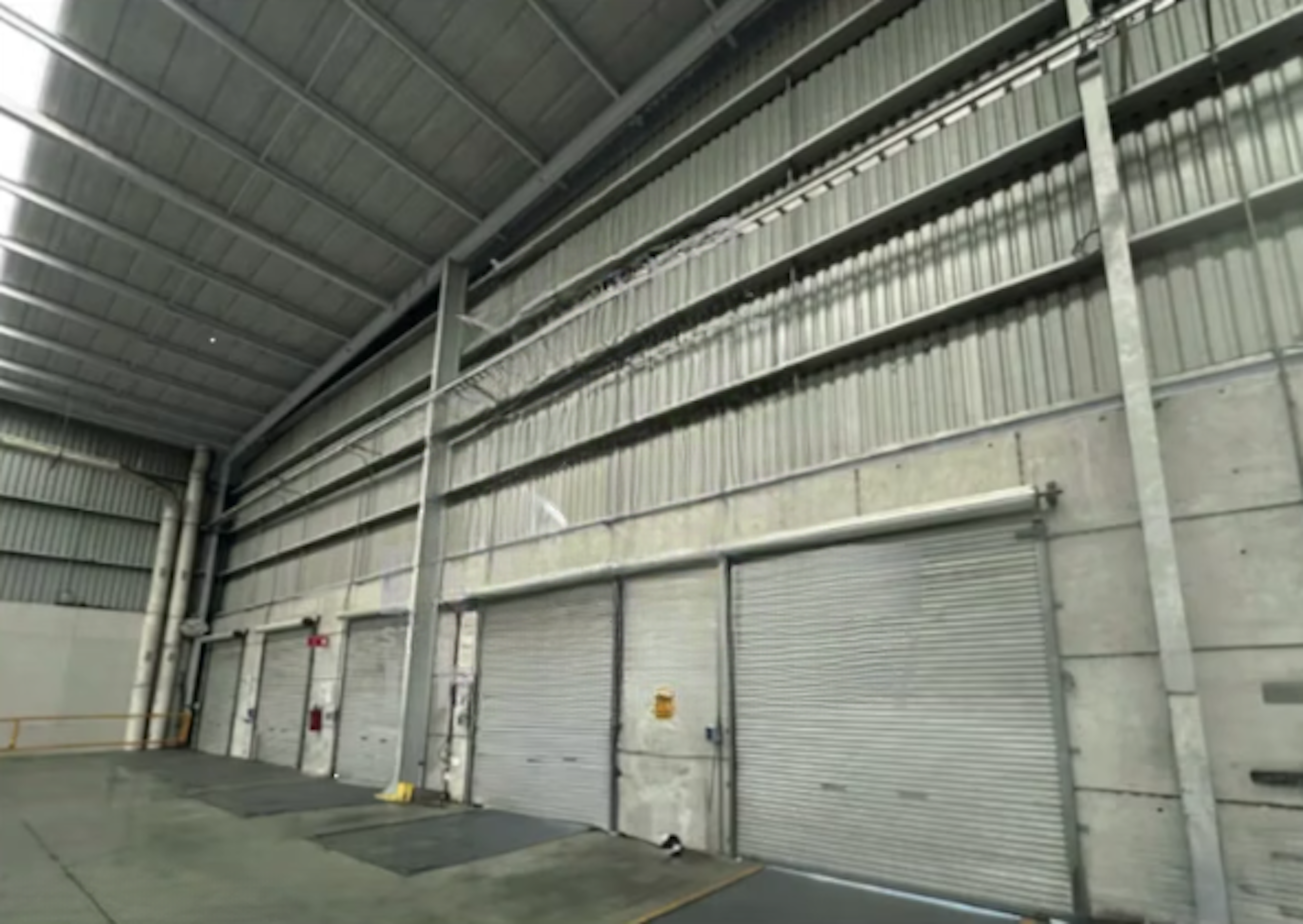 La imagen muestra el interior de una nave industrial o almacén de gran altura. Se observan paredes de concreto con revestimiento metálico corrugado en la parte superior. Hay varias puertas de garage metálicas enrollables a lo largo de la pared. El techo es alto con vigas y estructuras metálicas visibles. El espacio parece amplio y diseñado para almacenamiento o actividades industriales.