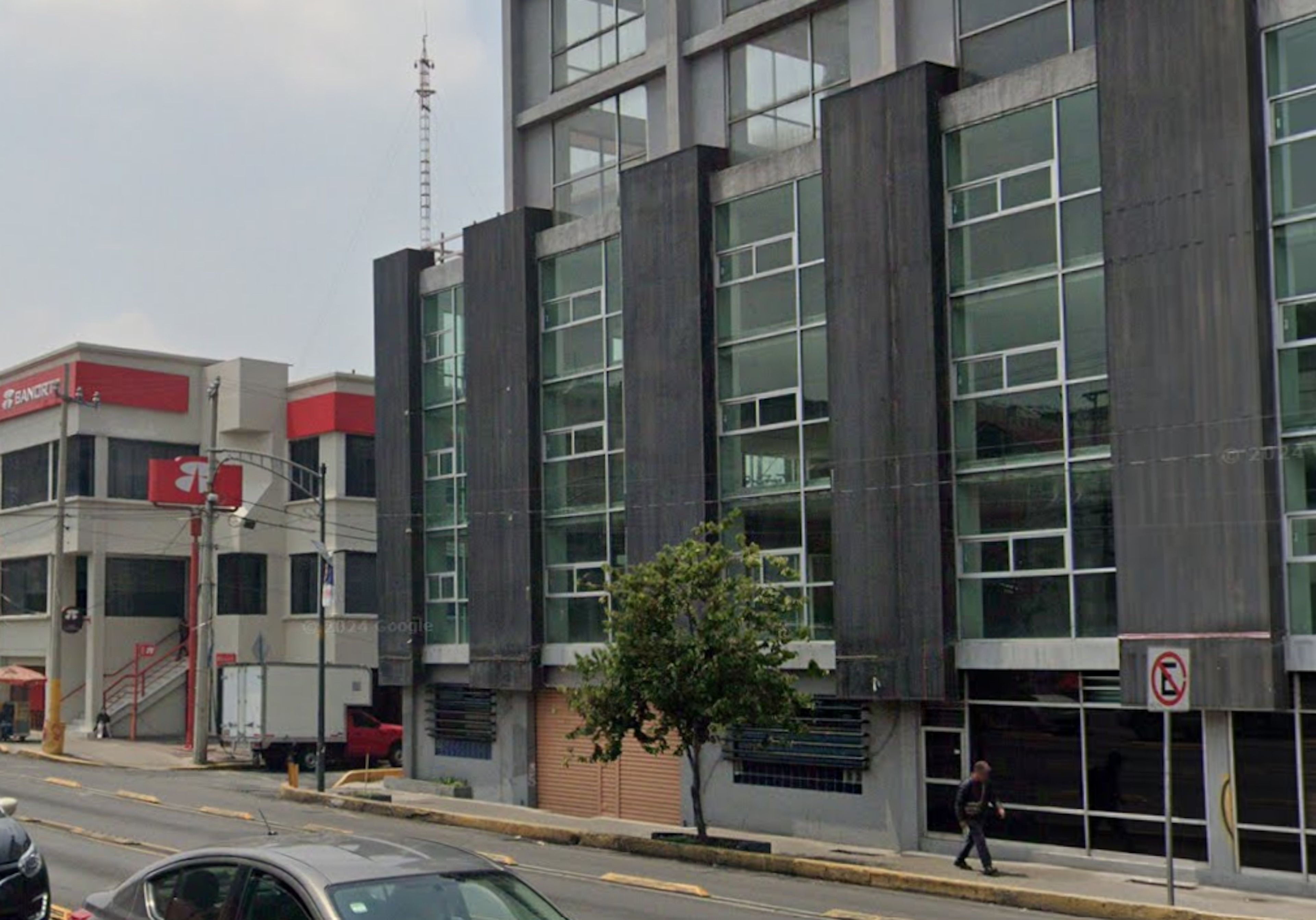 La imagen muestra un edificio moderno de varios pisos con fachada de vidrio y paneles oscuros. Tiene grandes ventanales y está ubicado en una zona urbana. A la izquierda se ve parte de otro edificio con colores rojo y blanco, posiblemente comercial. La calle tiene aceras, árboles y señales de tráfico. El estilo arquitectónico sugiere un uso mixto, posiblemente oficinas o apartamentos en los pisos superiores.