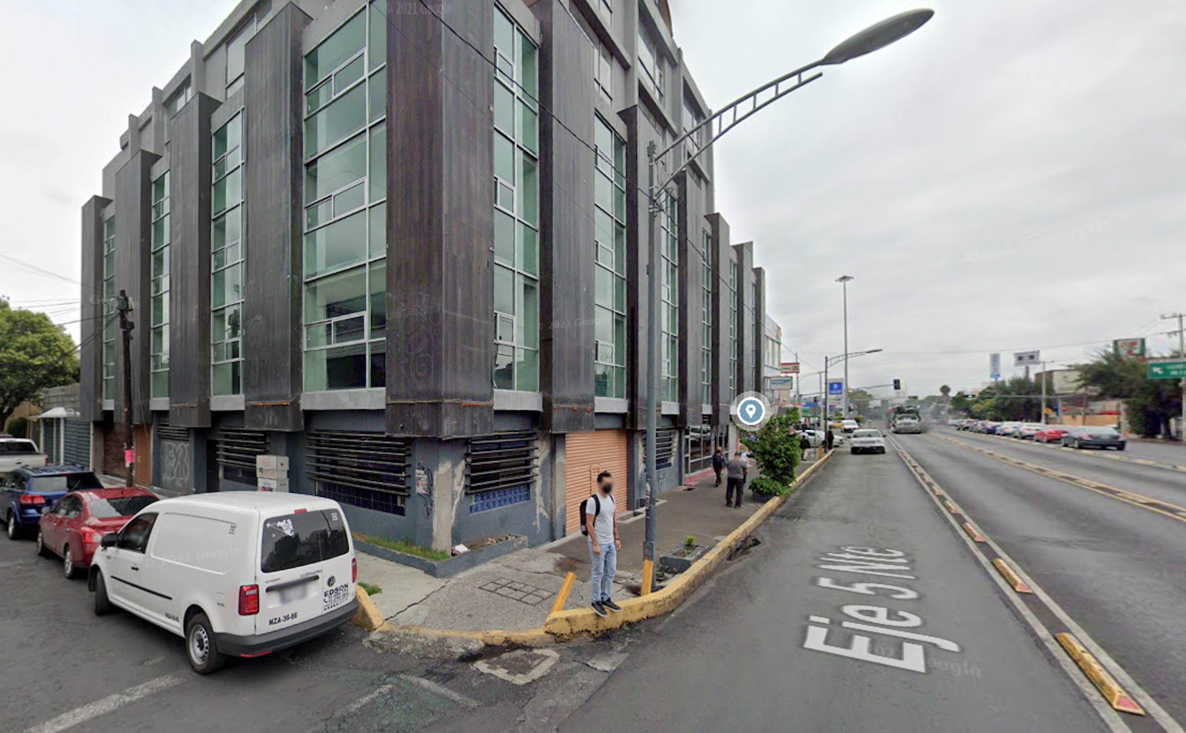 La imagen muestra un edificio moderno de varios pisos con fachada de vidrio y metal en una esquina urbana. Parece ser un inmueble comercial o de oficinas en una zona céntrica. La calle está pavimentada y hay vehículos estacionados cerca. Se observan algunos peatones y postes de luz. El entorno sugiere una área comercial o de negocios en desarrollo.