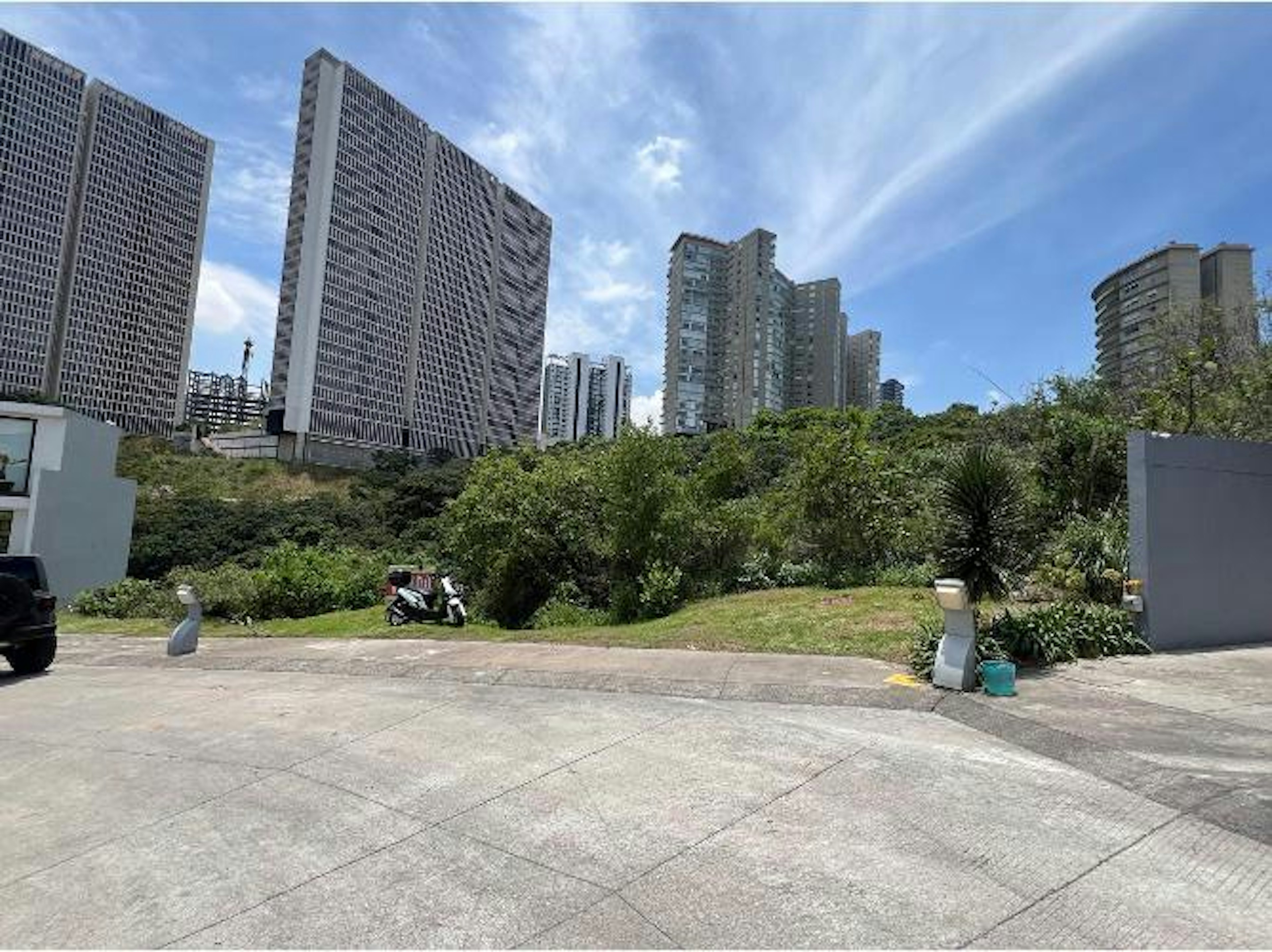 La imagen muestra un área urbana con altos edificios residenciales y de oficinas. En primer plano se ve un terreno con vegetación y algunas estructuras pequeñas. El cielo es azul con nubes dispersas. La escena sugiere una zona de desarrollo inmobiliario en una ciudad moderna, con una mezcla de espacios verdes y construcciones de gran altura. El contraste entre la naturaleza y los rascacielos es notable.