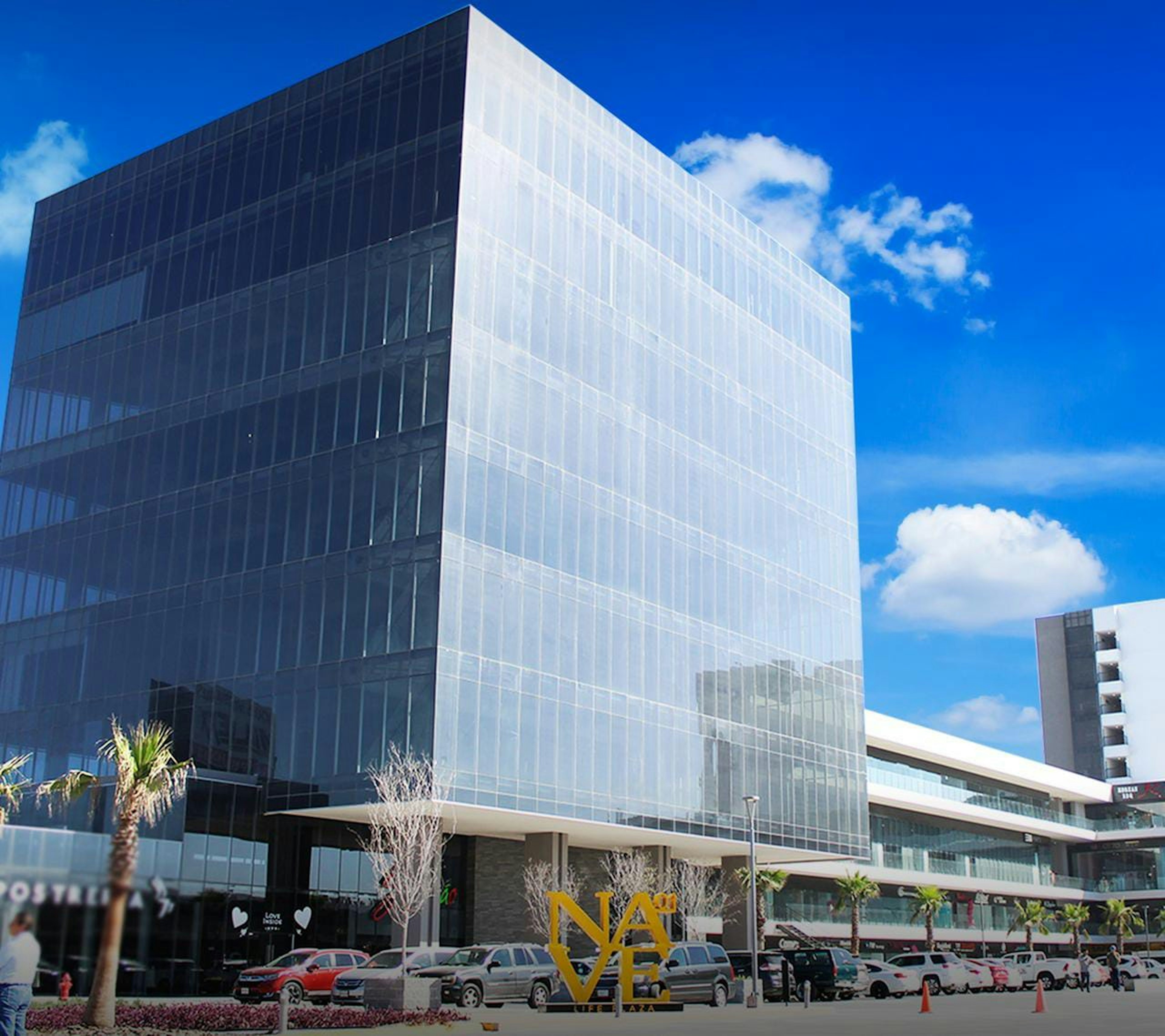 La imagen muestra un moderno edificio de oficinas con fachada de vidrio, parte oscura y parte clara, reflejando el cielo azul. En la planta baja hay un letrero que dice NAVE. El edificio está rodeado de palmeras, otros edificios y un estacionamiento con varios autos. La arquitectura es contemporánea y el ambiente parece ser de una zona comercial o de negocios en desarrollo.