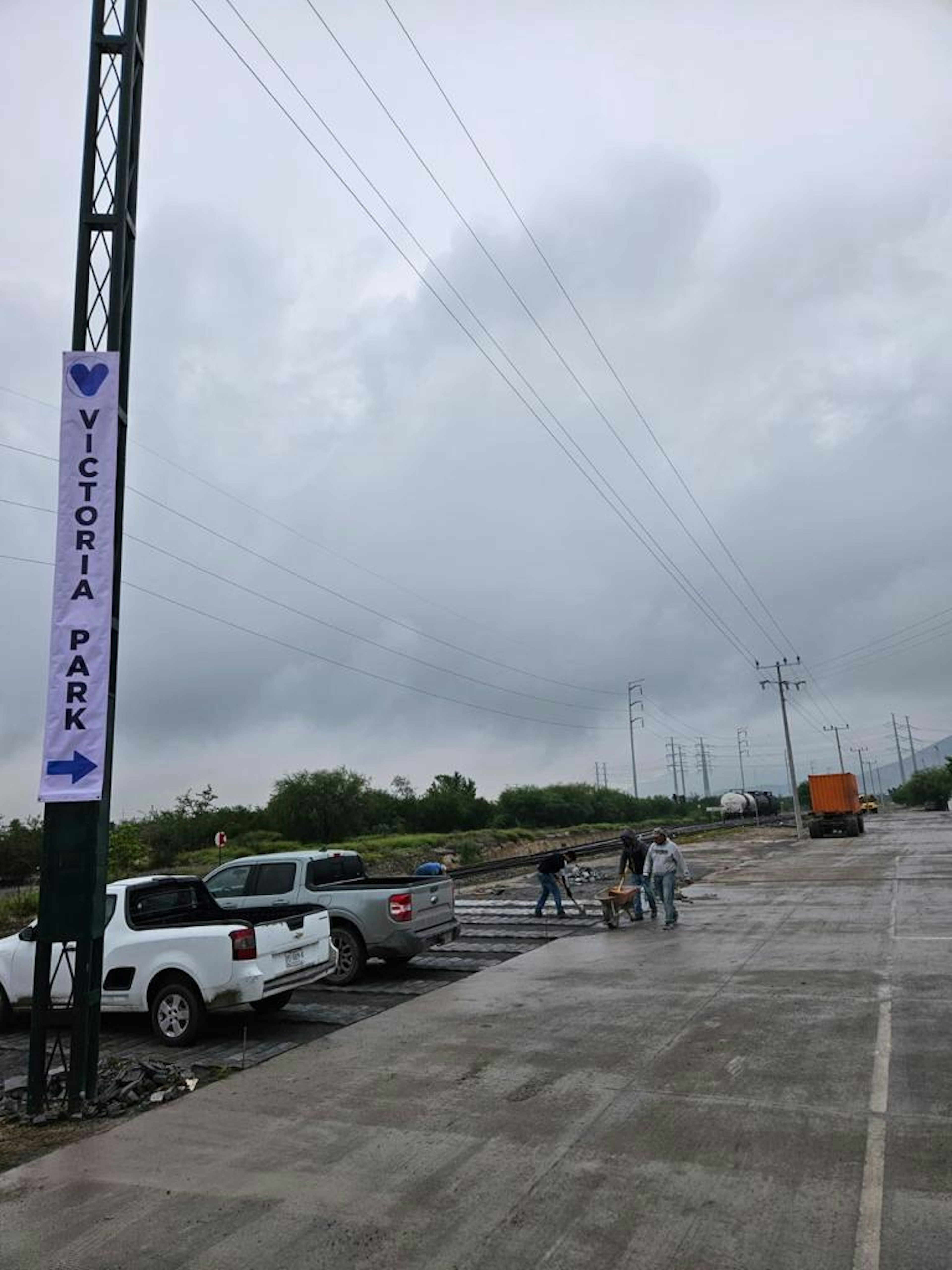 La imagen muestra un área industrial o en desarrollo con un cartel que indica Victoria Park. Se observan vehículos estacionados, trabajadores en el sitio, y una carretera en construcción. El cielo está nublado y hay líneas eléctricas visibles. El ambiente sugiere un proyecto de infraestructura o desarrollo inmobiliario en progreso.