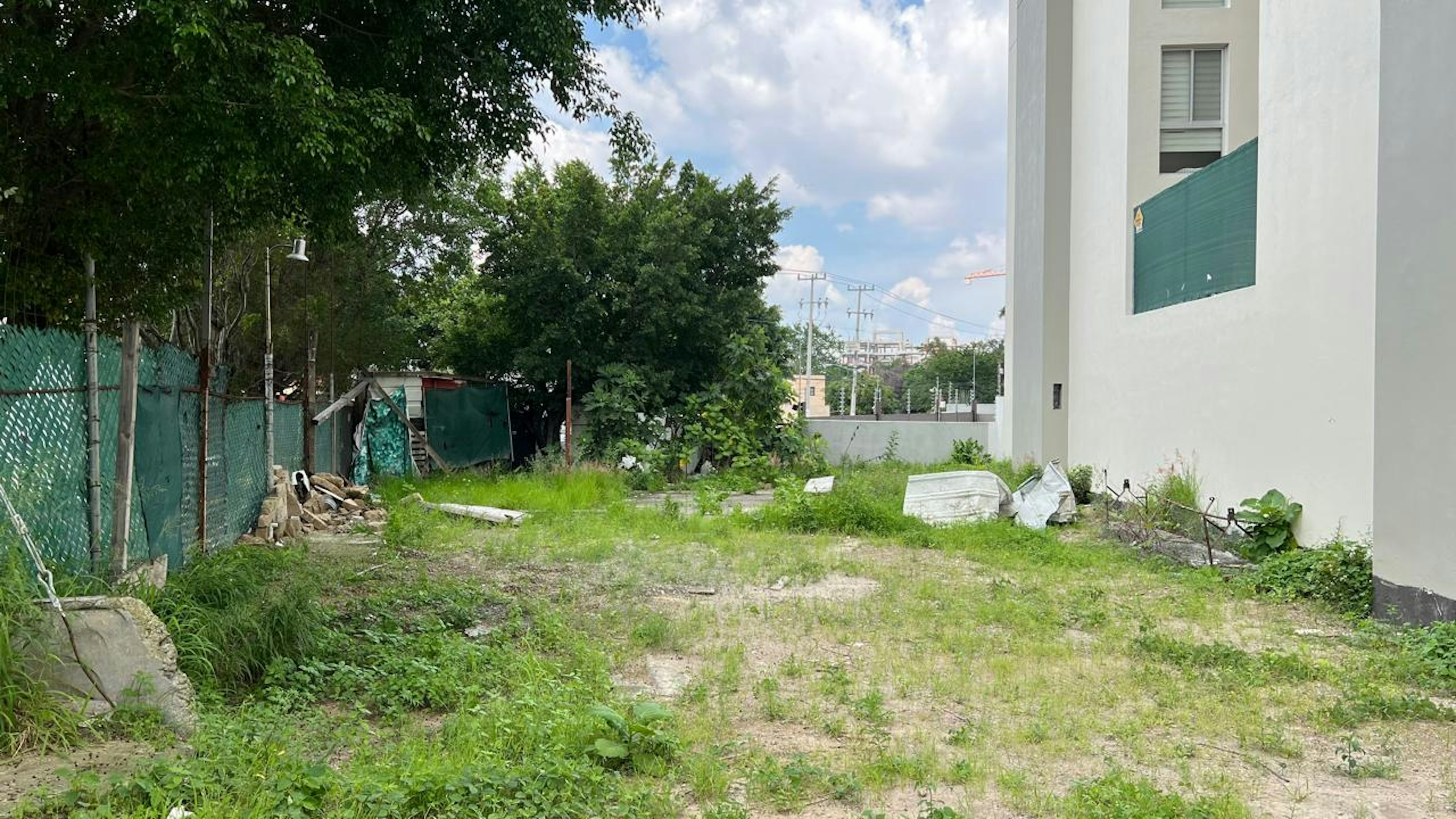 La imagen muestra un terreno baldío en una zona urbana. El lote está cubierto de hierba y maleza, con algunos escombros y materiales dispersos. Está rodeado por una valla verde y se ve un edificio blanco moderno a un lado. Al fondo se aprecian árboles y postes eléctricos, sugiriendo una ubicación en desarrollo con potencial para construcción o mejora inmobiliaria.