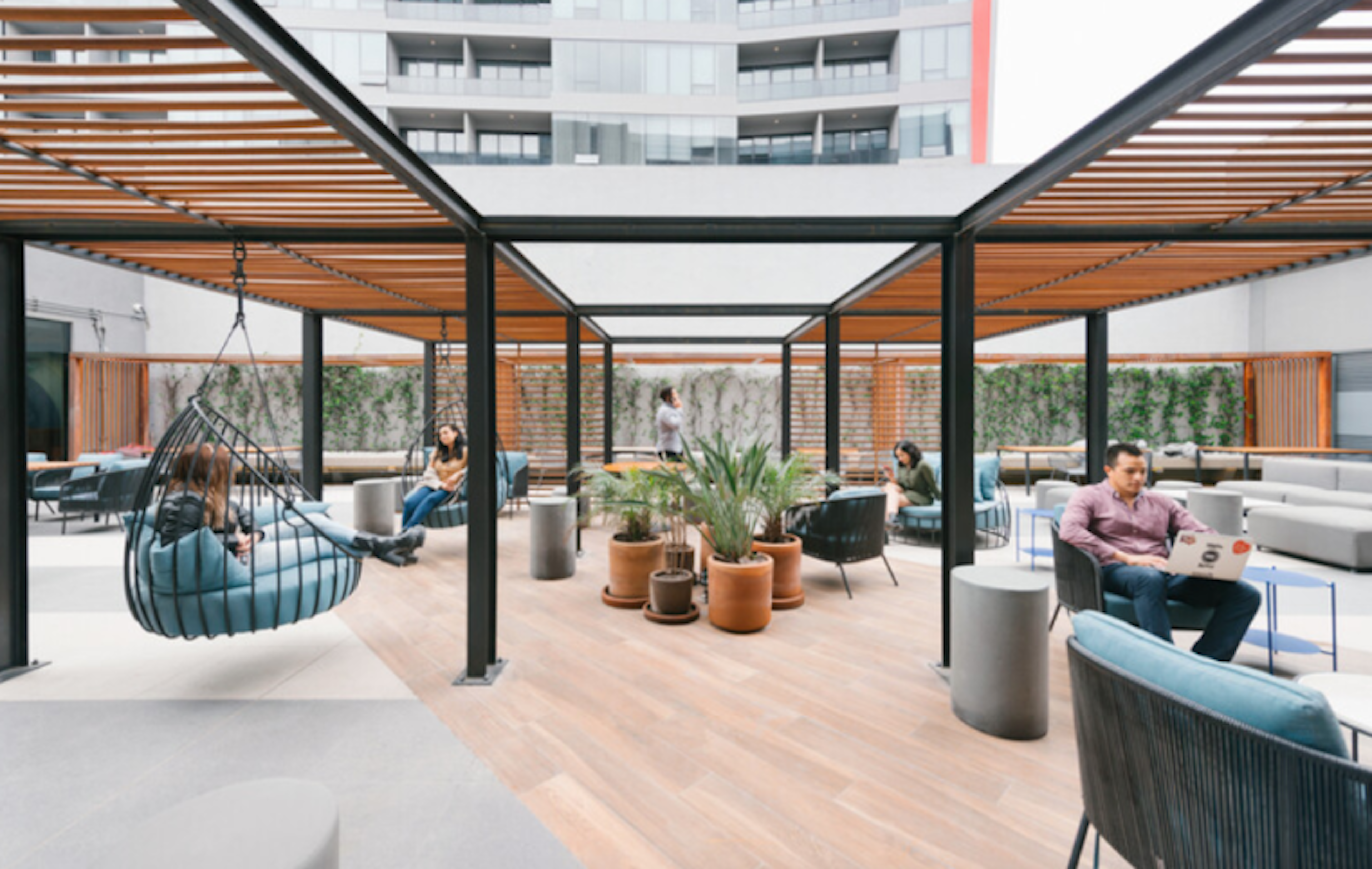 La imagen muestra un moderno espacio de coworking al aire libre. Cuenta con una estructura de techo con listones de madera, áreas de asiento variadas incluyendo sillas colgantes y sofás. El espacio está decorado con plantas en macetas y tiene un suelo de madera. Varias personas están utilizando el área para trabajar o relajarse. El diseño combina elementos naturales con un estilo contemporáneo, creando un ambiente acogedor y funcional para el trabajo flexible.