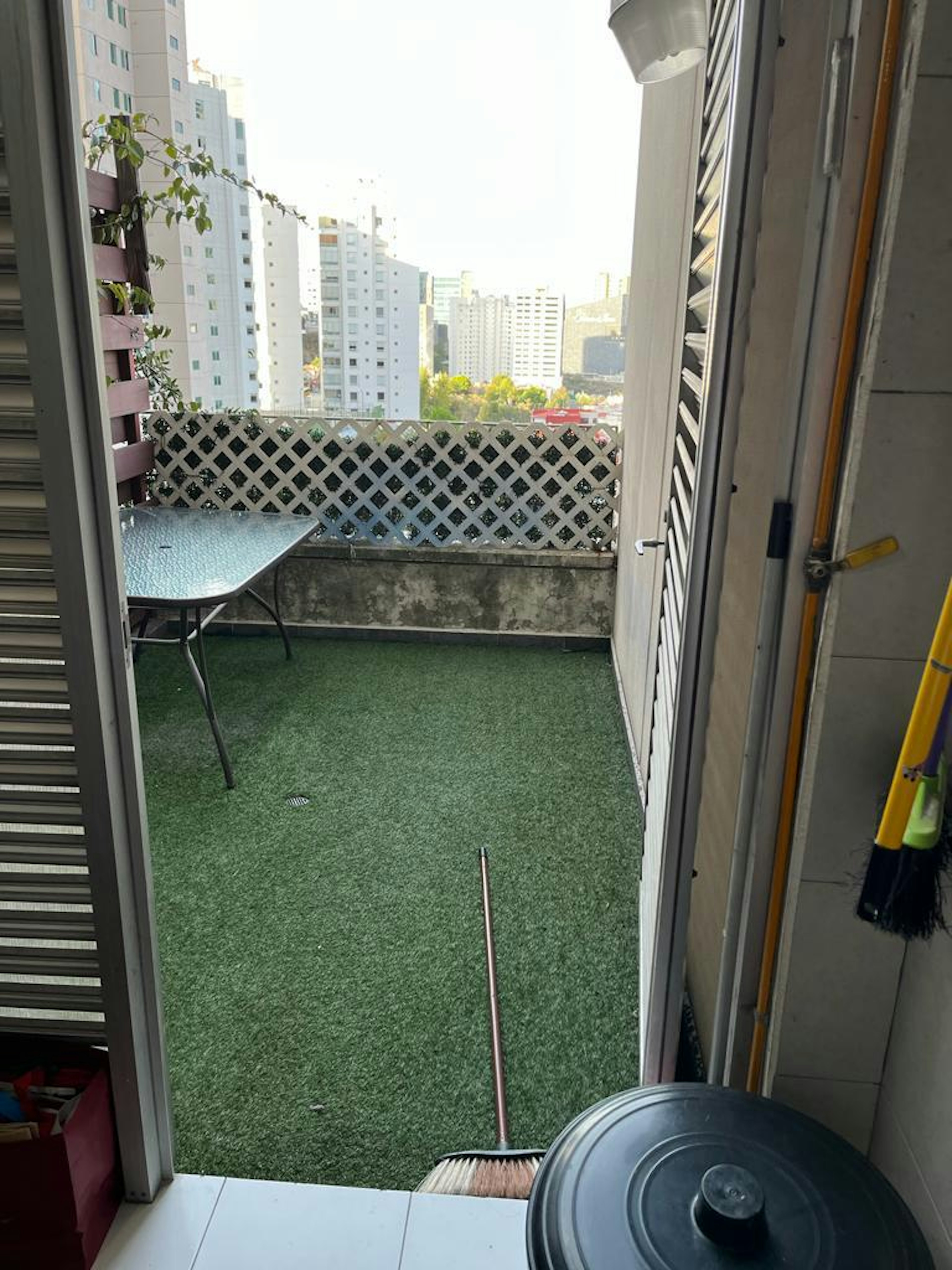 La imagen muestra un pequeño balcón o terraza de un apartamento en una zona urbana densamente poblada. El espacio está cubierto con césped artificial y tiene una mesa plegable. Una rejilla de madera sirve como baranda, ofreciendo privacidad y vistas parciales de los edificios de gran altura circundantes. Se pueden ver elementos de limpieza como una escoba, lo que sugiere un espacio funcional y bien mantenido.