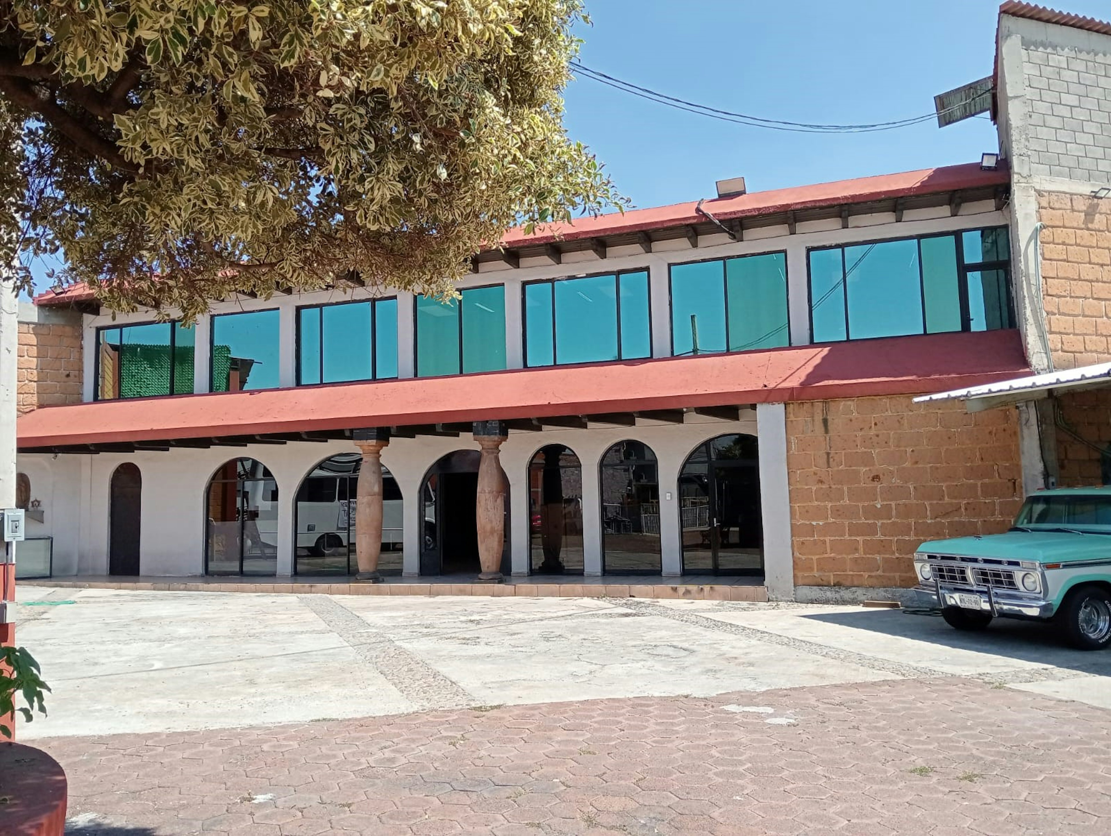 La imagen muestra un edificio de estilo colonial con arcos y columnas en la planta baja. La parte superior tiene grandes ventanas de cristal tintado verde. El techo es rojo y hay un árbol frondoso a la izquierda. El edificio parece ser un local comercial o de oficinas, con una plaza pavimentada en frente. Un coche clásico azul está estacionado a la derecha, dando un toque nostálgico a la escena.