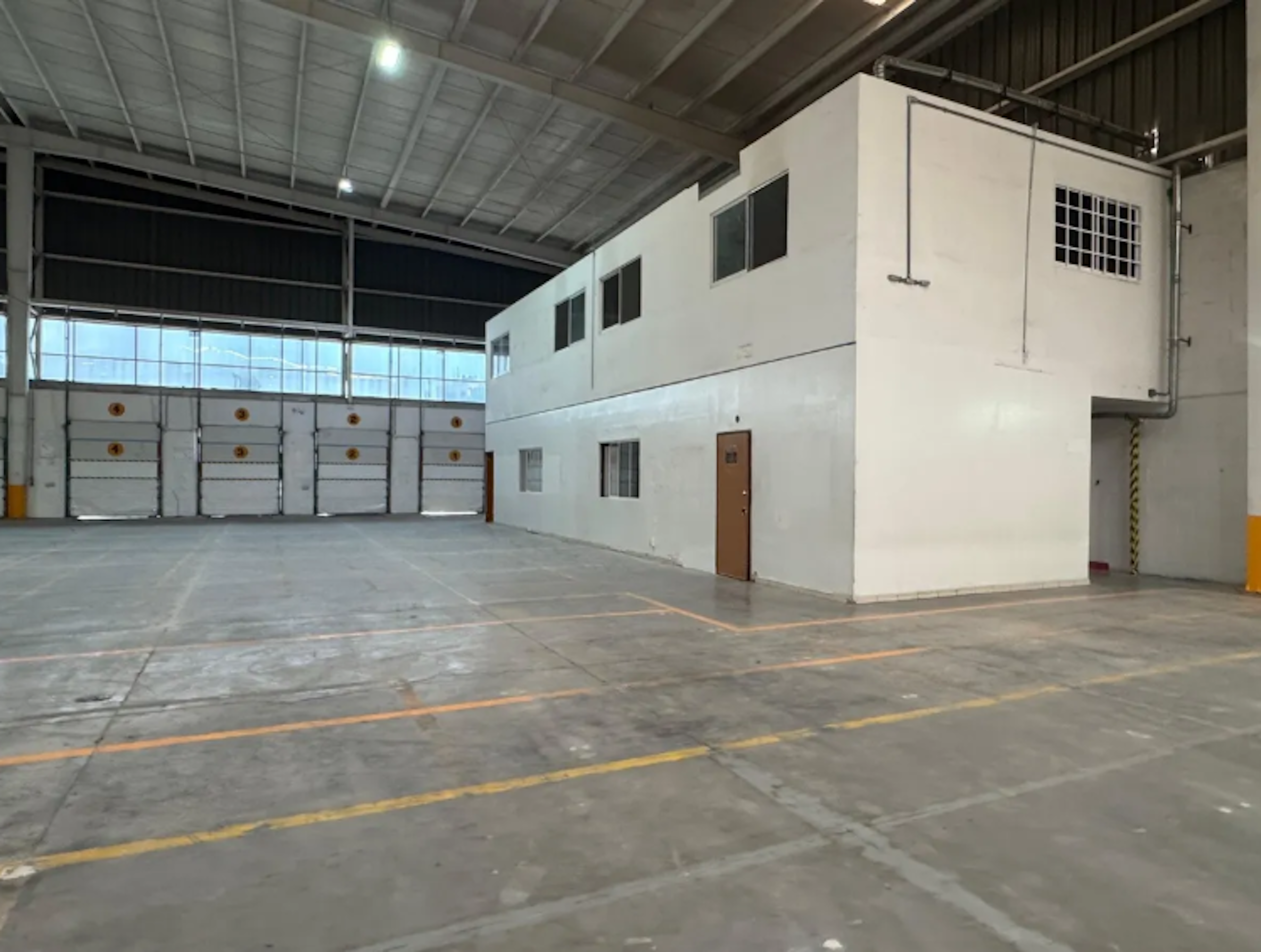 La imagen muestra el interior de una bodega industrial moderna. Se observa un amplio espacio con piso de concreto y techo alto. A la izquierda hay una serie de puertas de carga, y a la derecha se encuentra una estructura de dos pisos en color blanco con ventanas y una puerta de acceso. El espacio parece estar bien iluminado y organizado, ideal para almacenamiento o operaciones logísticas.