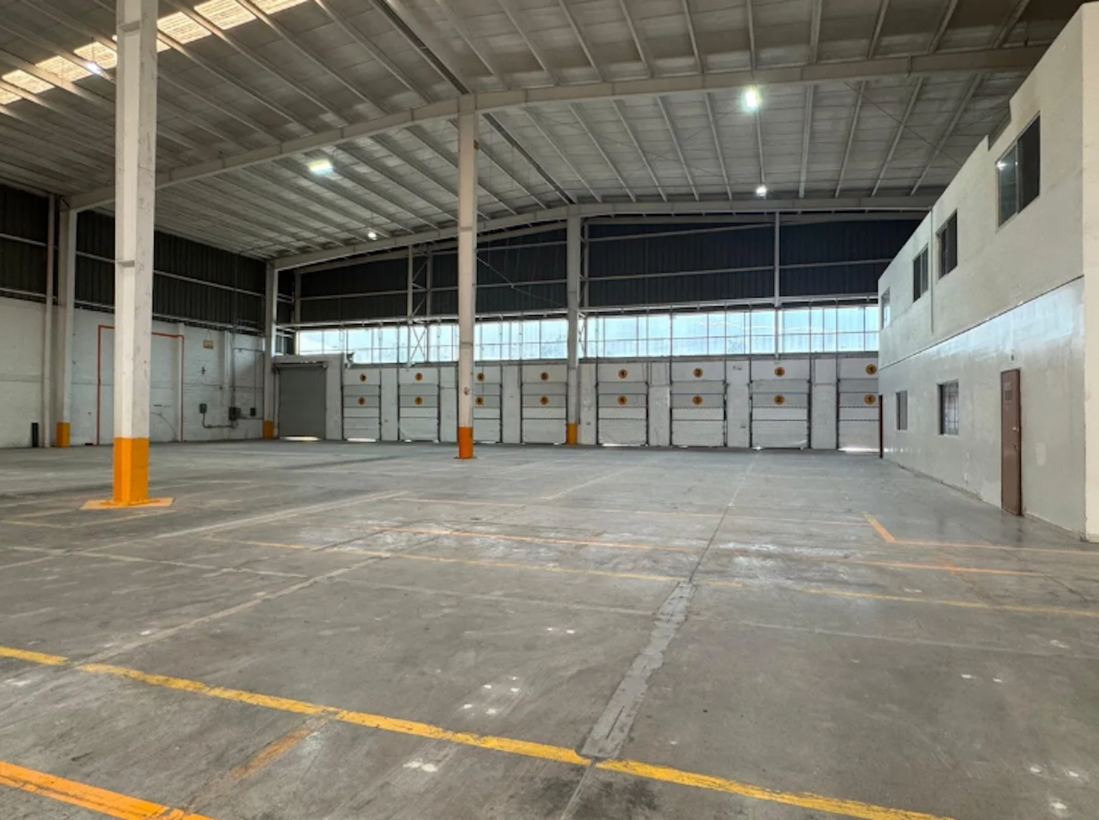 La imagen muestra un amplio almacén o nave industrial con techo alto y estructura metálica. El espacio está vacío, con suelo de concreto y líneas amarillas pintadas. A lo largo de una pared hay una serie de puertas de carga blancas con marcas naranjas. El edificio tiene ventanas altas que permiten la entrada de luz natural. Las columnas y parte inferior de las paredes están pintadas de naranja, dando un toque de color al espacio industrial.