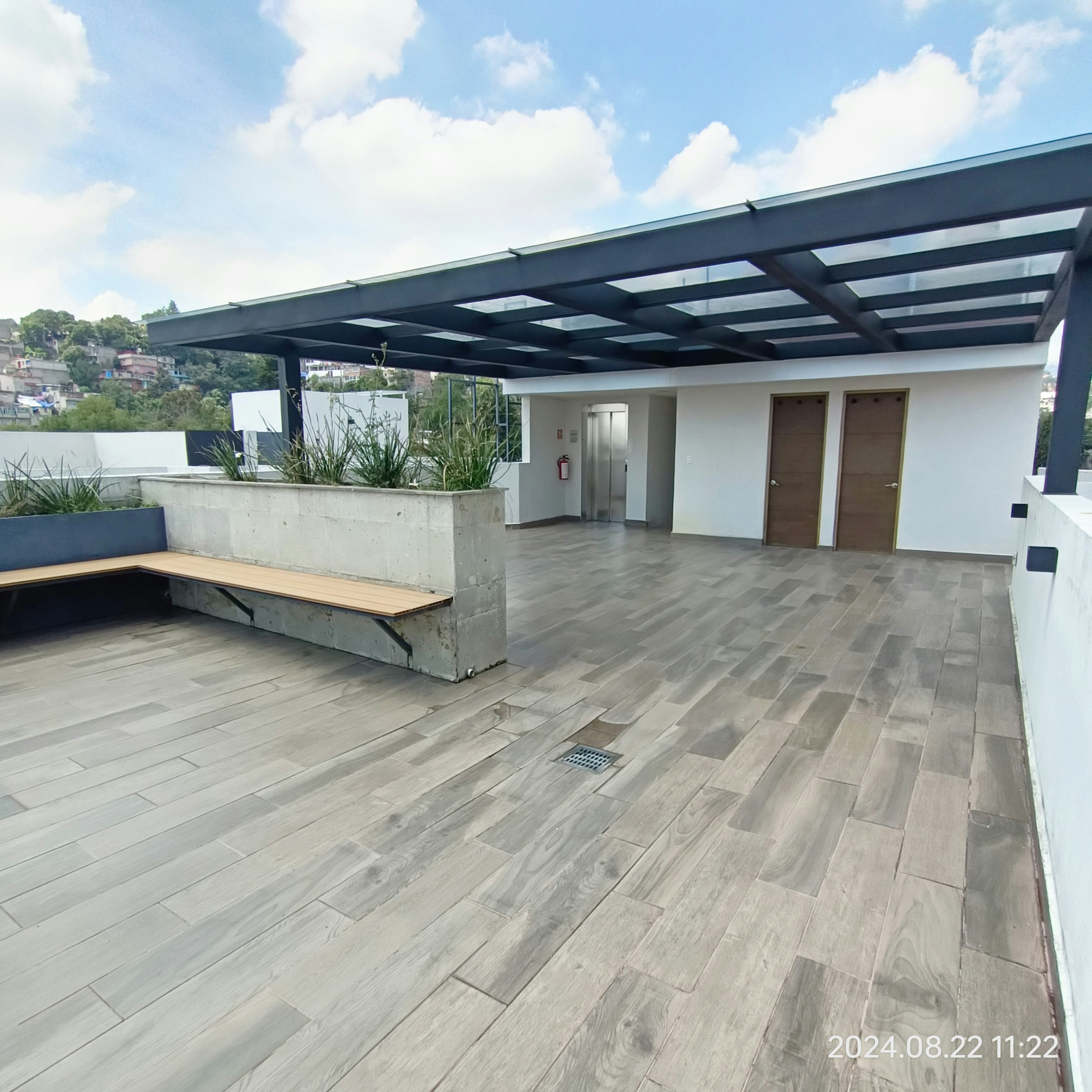 La imagen muestra una terraza moderna en la azotea de un edificio. Cuenta con un piso de madera amplio, una pérgola metálica que proporciona sombra, y jardineras de concreto con plantas. Se observan puertas que conducen al interior, un banco integrado y vistas a un área residencial en la distancia. El diseño es minimalista y funcional, ideal para espacios al aire libre en desarrollos inmobiliarios contemporáneos.