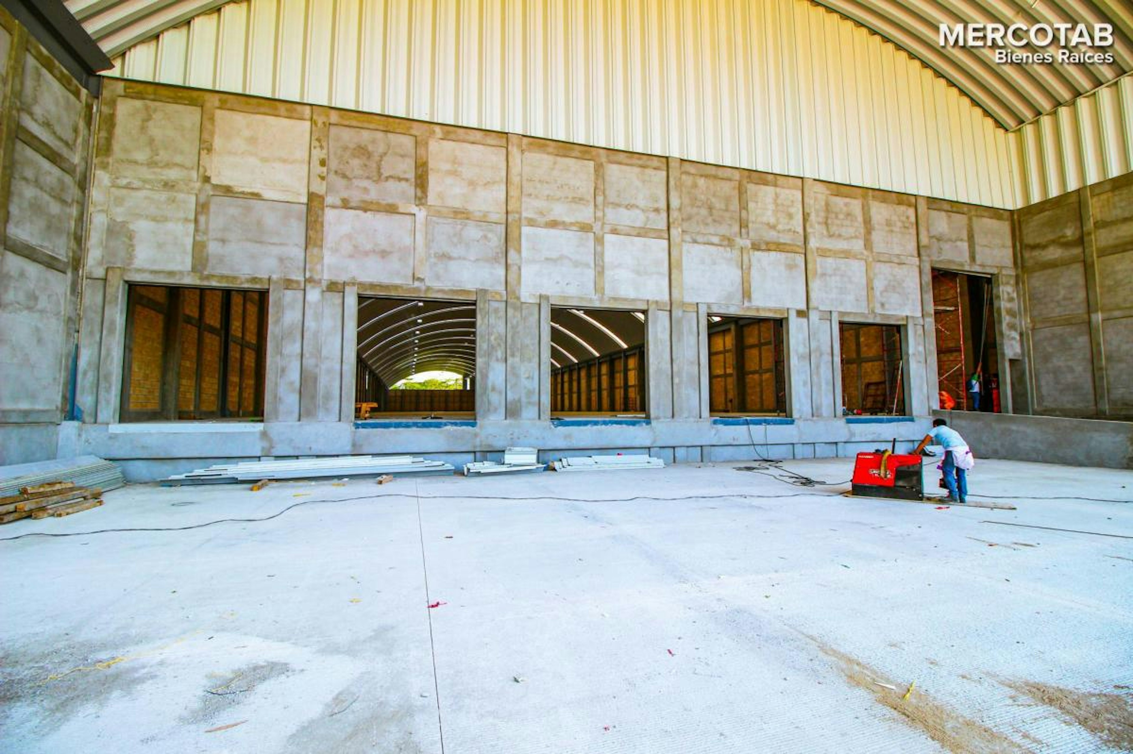 La imagen muestra una construcción en proceso de un gran espacio industrial o comercial. Se observa una estructura con techo arqueado, paredes de concreto con aberturas para ventanas y puertas. El piso es de cemento y hay materiales de construcción dispersos. Se aprecia una persona trabajando, lo que indica que la obra está en progreso. El logo MERCOTAB Bienes Raíces sugiere que es un proyecto inmobiliario comercial.