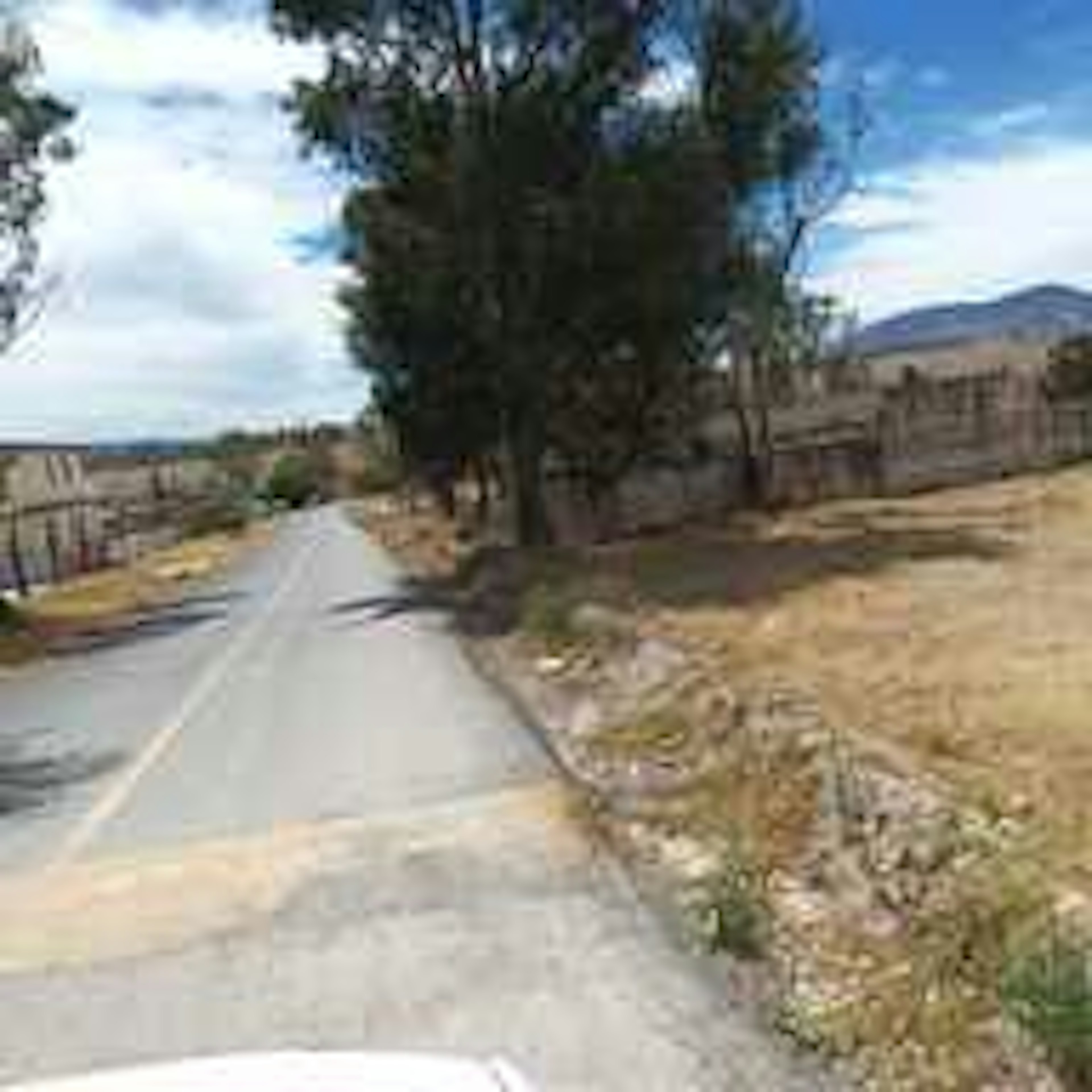 La imagen muestra una carretera rural estrecha que atraviesa un paisaje árido. A los lados hay árboles dispersos y vegetación seca. Se observan montañas en el fondo y un cielo parcialmente nublado. El terreno parece ser bastante seco y poco desarrollado, típico de una zona rural o suburbana con poca infraestructura. Este tipo de ubicación podría ser atractiva para desarrollos inmobiliarios de bajo costo o propiedades rurales.