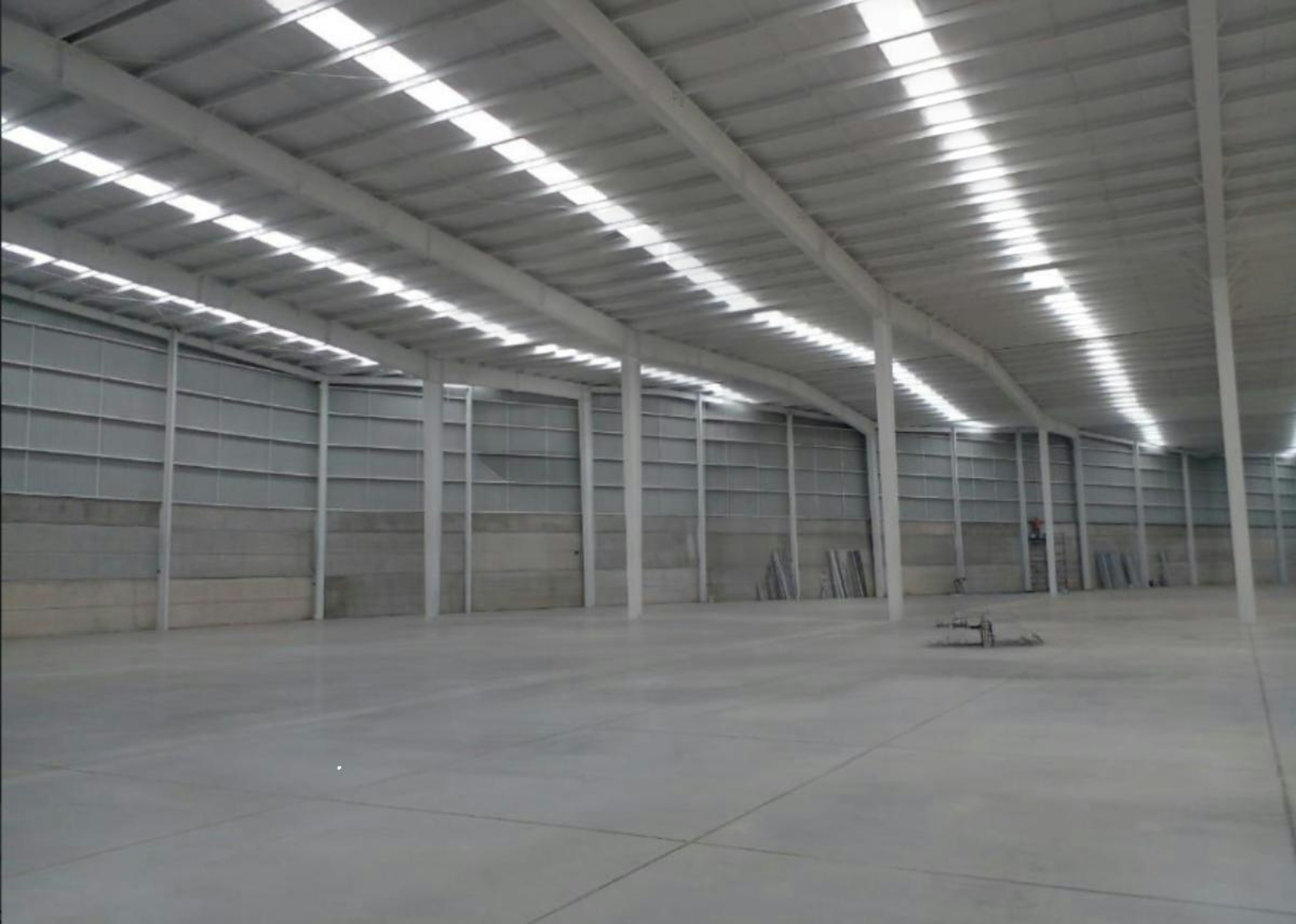 La imagen muestra un amplio almacén o nave industrial con techo alto y luminoso. Las paredes son de concreto gris y el piso es de cemento pulido. Se observan columnas metálicas que sostienen el techo. Al fondo hay puertas grandes, posiblemente para carga y descarga. En primer plano se ve un logo y texto que dice Strategic Industry Logistics. El espacio está prácticamente vacío, sugiriendo que está listo para ser utilizado o alquilado.