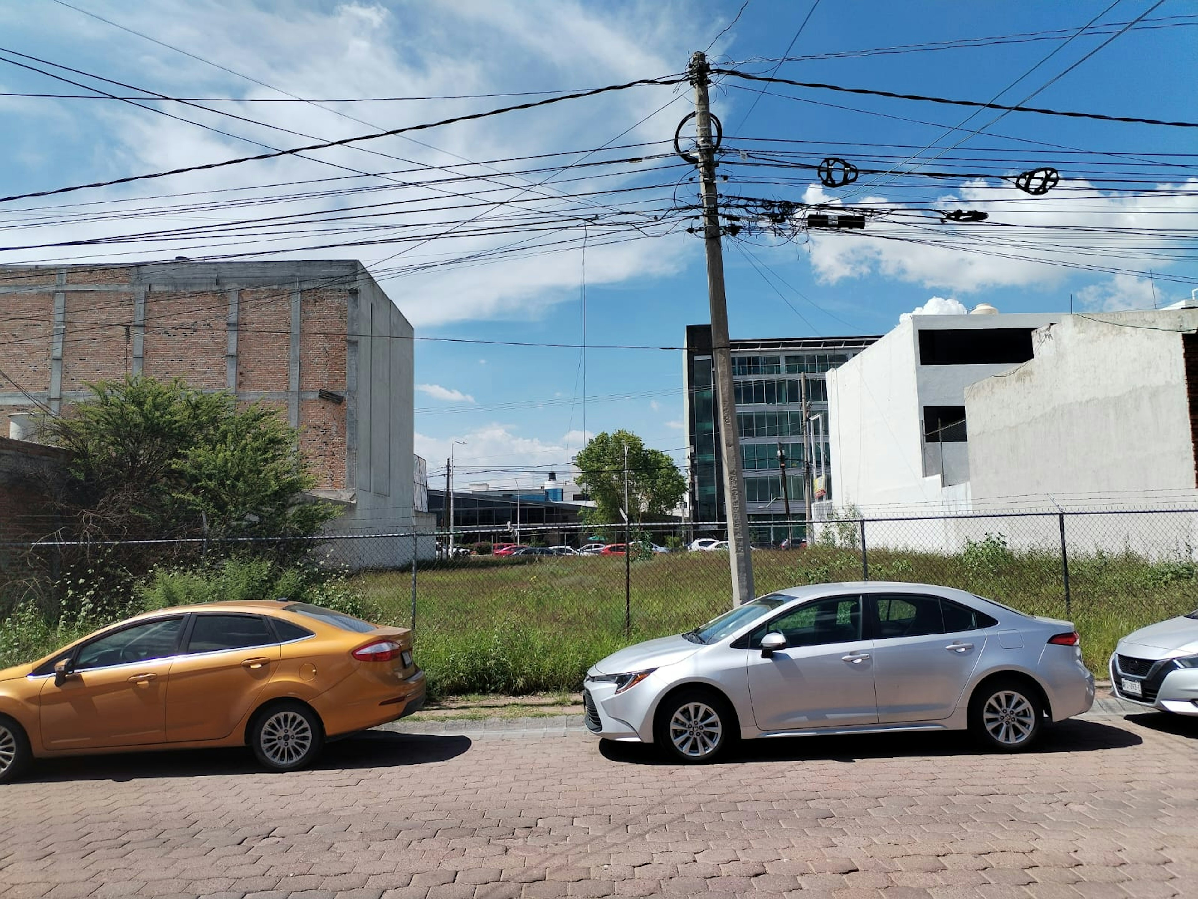 La imagen muestra un área urbana con edificios de ladrillo y concreto, algunos en construcción o abandonados. Hay un terreno baldío cercado con vegetación silvestre. Se observan varios autos estacionados en primer plano sobre una calle pavimentada. Numerosos cables eléctricos cruzan el cielo, conectados a postes. El entorno sugiere una zona en desarrollo o transición, con potencial para proyectos inmobiliarios.