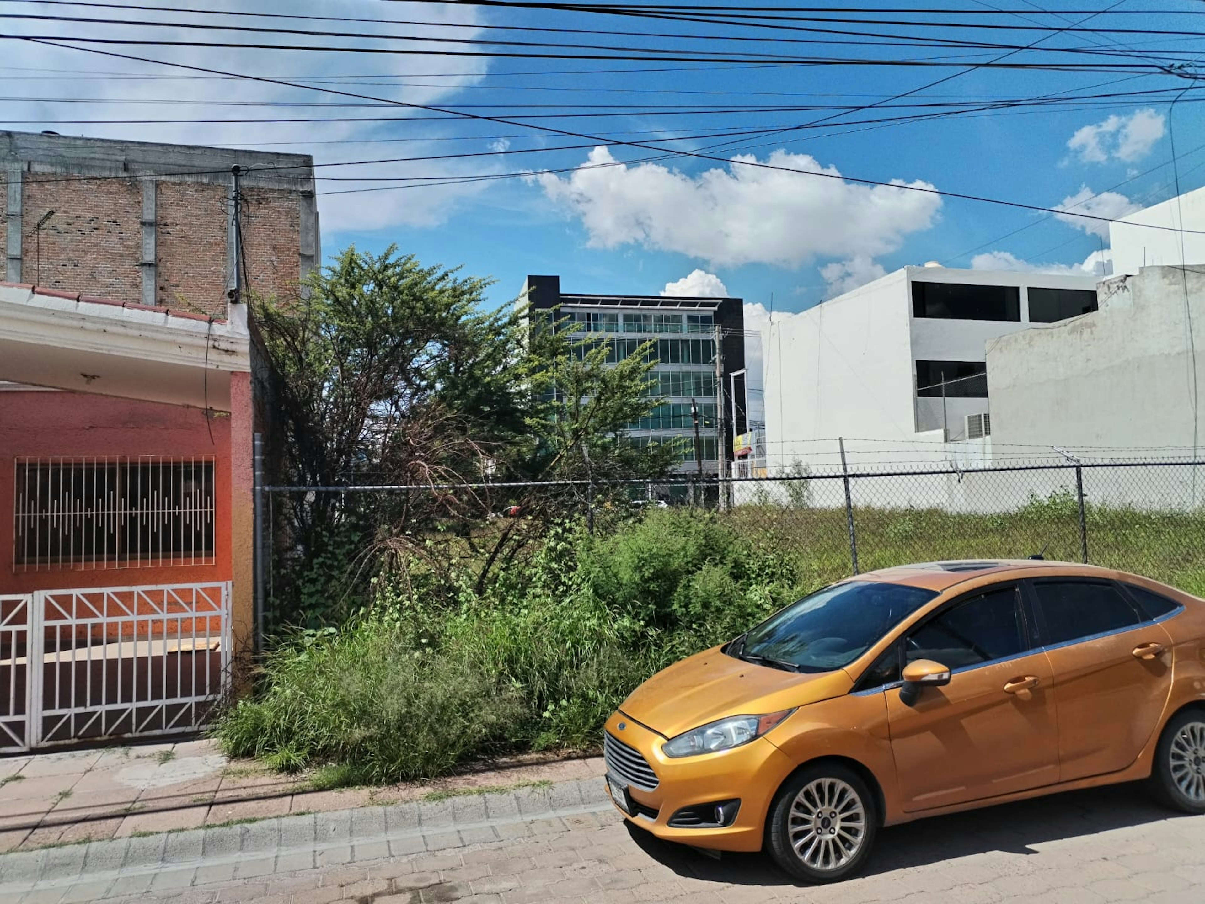 La imagen muestra una escena urbana con diversos elementos inmobiliarios. Se ve un edificio moderno de varios pisos con fachada de vidrio al fondo, junto a construcciones más antiguas de ladrillo y concreto. Hay un terreno baldío con vegetación silvestre, delimitado por una cerca. En primer plano se observa parte de una casa con reja y un auto amarillo estacionado en la calle. Los cables eléctricos atraviesan el cielo azul.