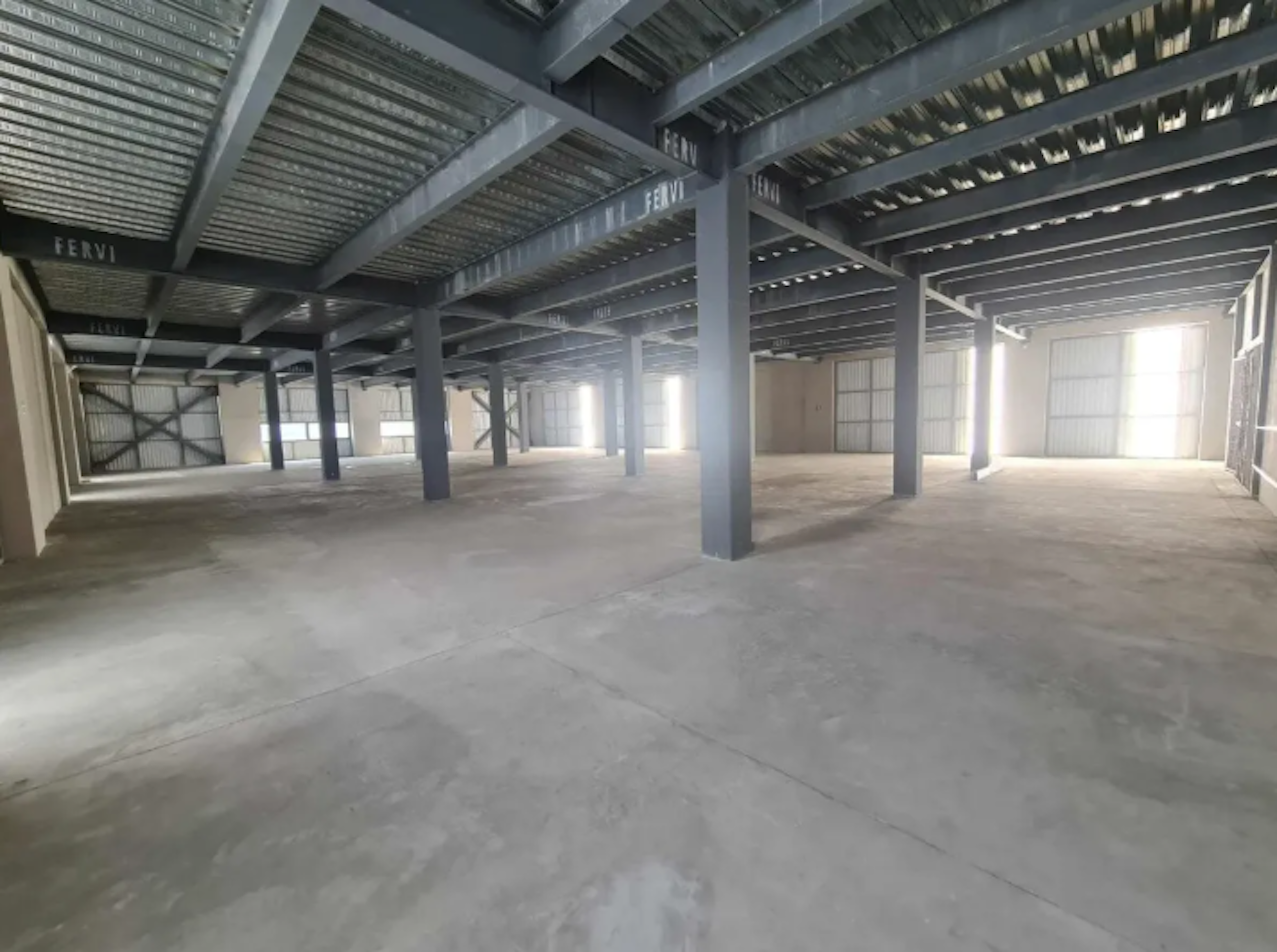 La imagen muestra un amplio espacio industrial o comercial vacío. Tiene un techo alto con vigas metálicas expuestas y columnas que sostienen la estructura. El piso es de concreto y las paredes laterales son claras. Hay varias entradas de luz natural, creando un ambiente luminoso. El espacio parece estar en buenas condiciones y listo para ser utilizado, ideal para almacenamiento, producción o como espacio comercial de gran tamaño.