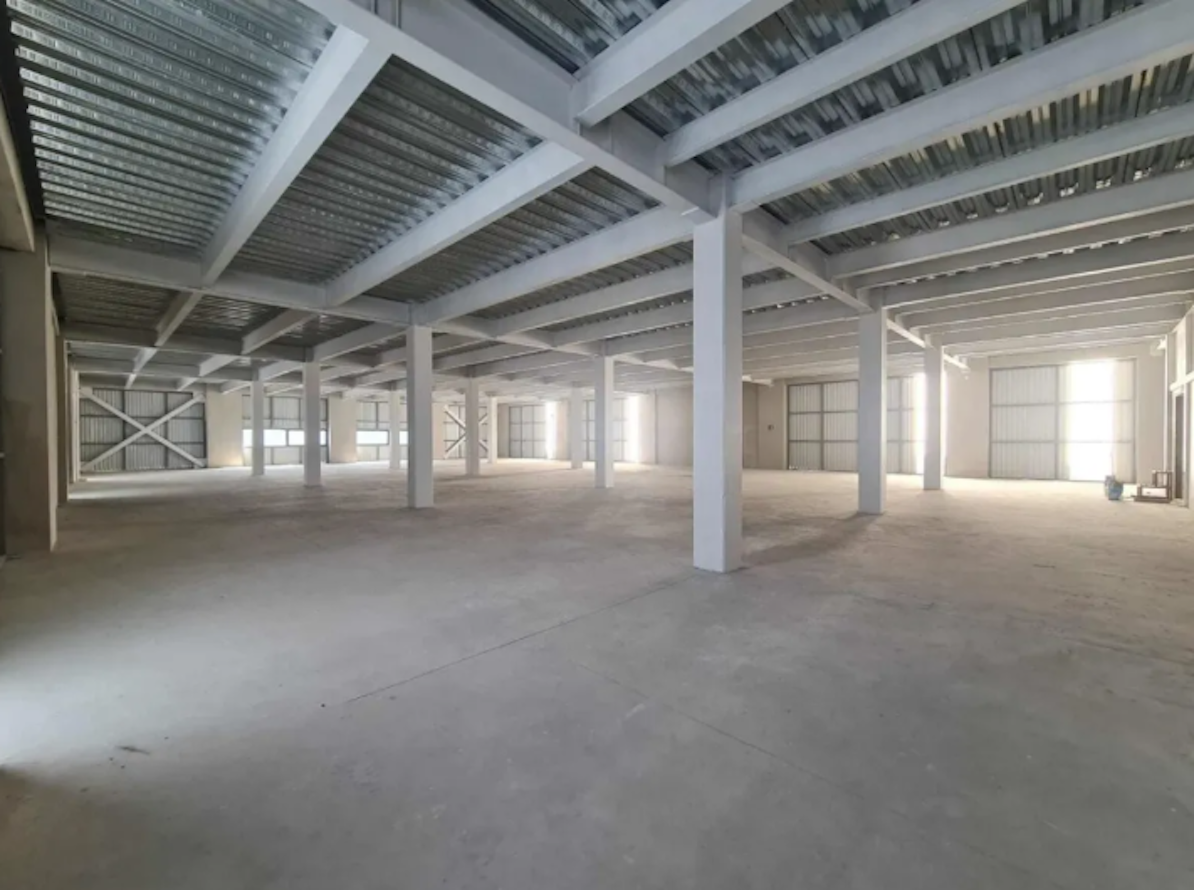 La imagen muestra un amplio espacio industrial o comercial vacío. Tiene un techo alto con vigas y columnas blancas que sostienen una estructura metálica. El piso es de concreto y hay ventanas grandes que permiten la entrada de luz natural. Este espacio parece estar en construcción o recién terminado, listo para ser adaptado según las necesidades del inquilino. Es ideal para almacenes, fábricas o espacios comerciales grandes.