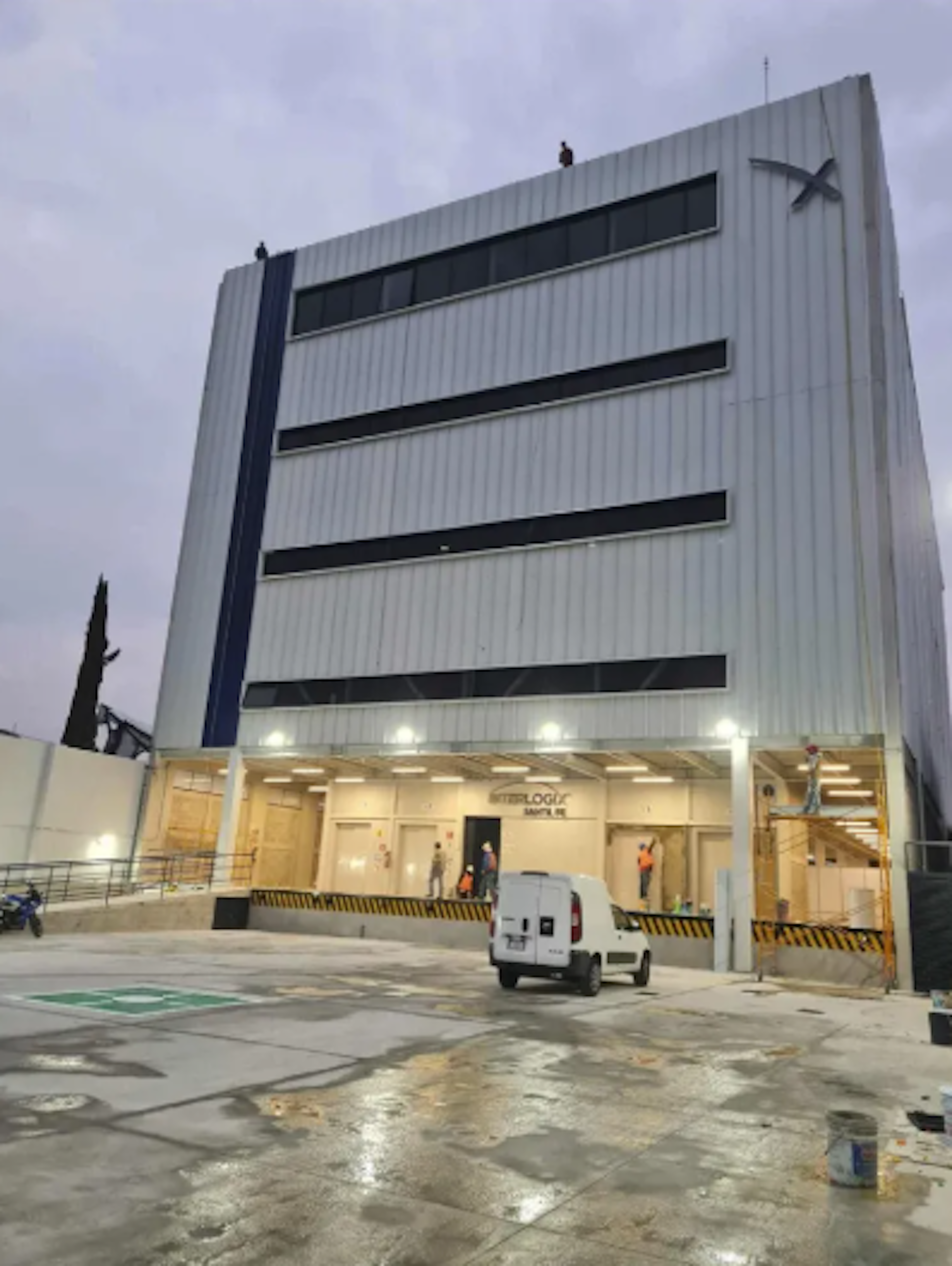 La imagen muestra un edificio industrial moderno de varios pisos con fachada blanca y ventanas oscuras. En la planta baja hay un área de carga iluminada con trabajadores y un vehículo. El edificio parece ser un centro logístico o almacén en construcción o renovación, con detalles como señalización de seguridad y equipos de construcción visibles. La estructura destaca por su diseño funcional y eficiente para operaciones logísticas.