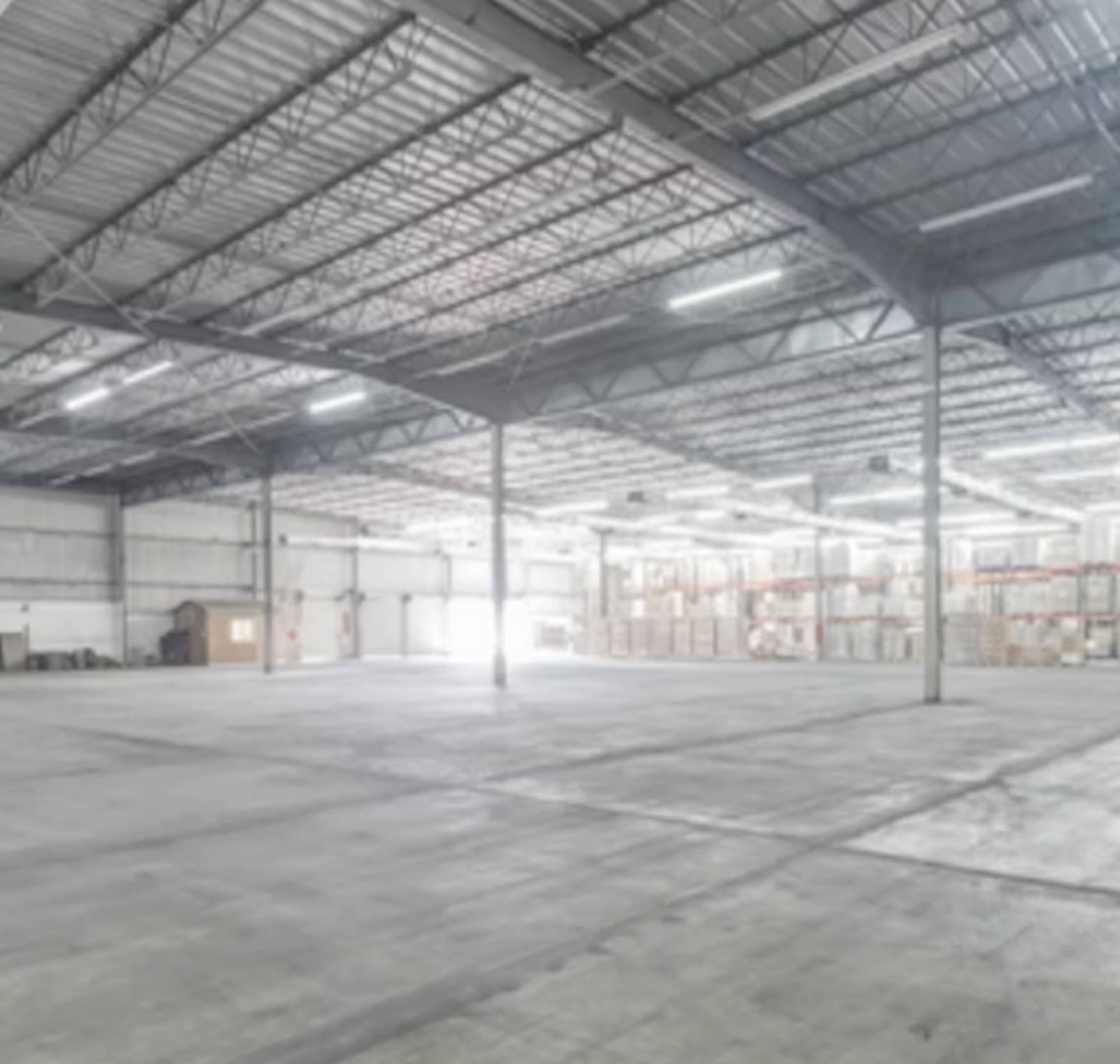 La imagen muestra un amplio almacén o nave industrial con techo alto de estructura metálica. El espacio está prácticamente vacío, con un piso de concreto y paredes blancas. Al fondo se pueden ver estanterías con cajas almacenadas. La iluminación proviene de lámparas fluorescentes en el techo. El espacio parece ideal para almacenamiento o distribución a gran escala, con mucha capacidad para equipos y mercancías.