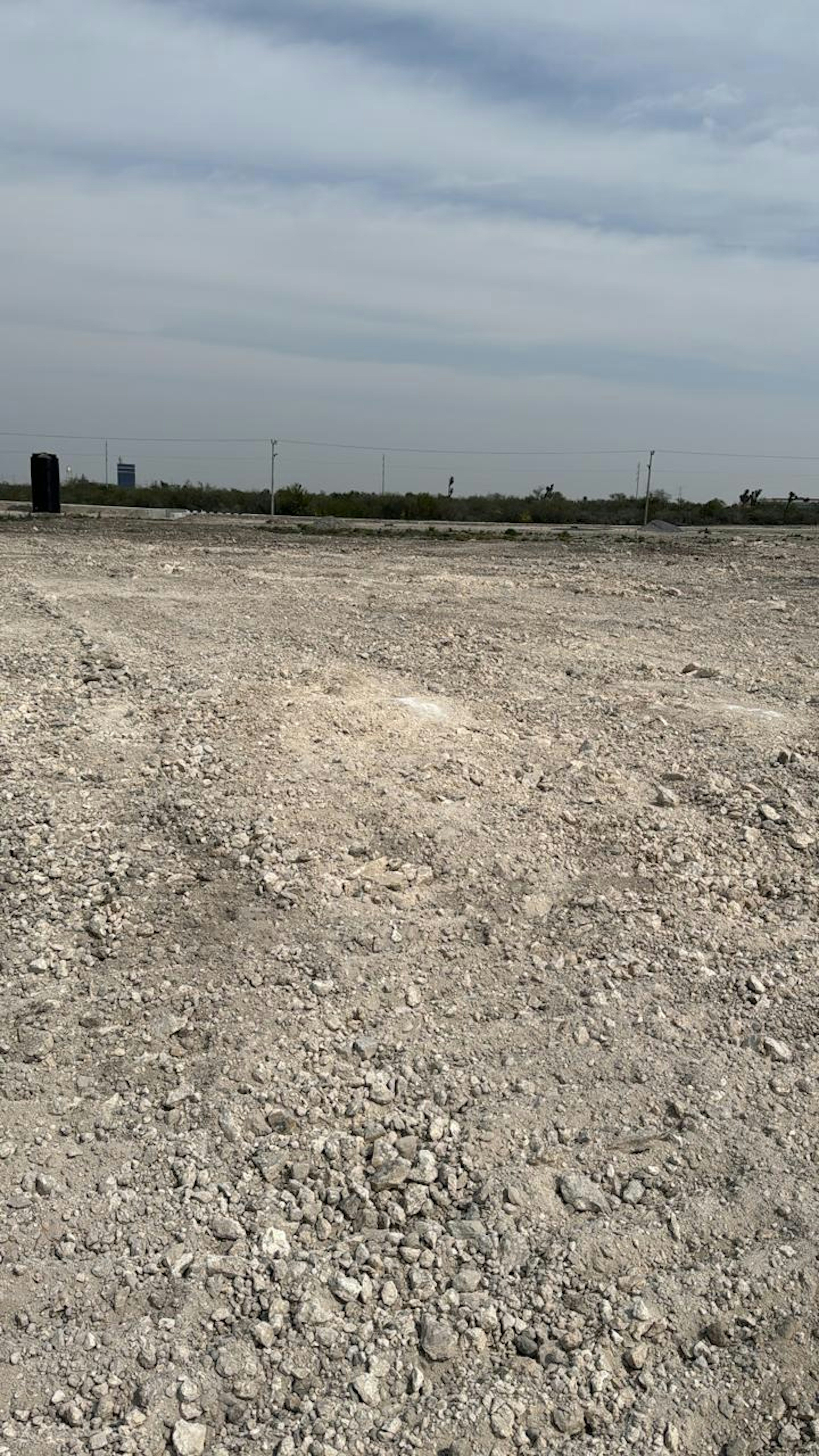 La imagen muestra un terreno baldío y árido, típico de una zona en desarrollo para proyectos inmobiliarios. El suelo es rocoso y polvoriento, sin vegetación visible. En el horizonte se observan postes eléctricos y una línea de árboles distantes, sugiriendo que el área está preparada para futuras construcciones. El cielo nublado añade un ambiente de anticipación al potencial desarrollo.