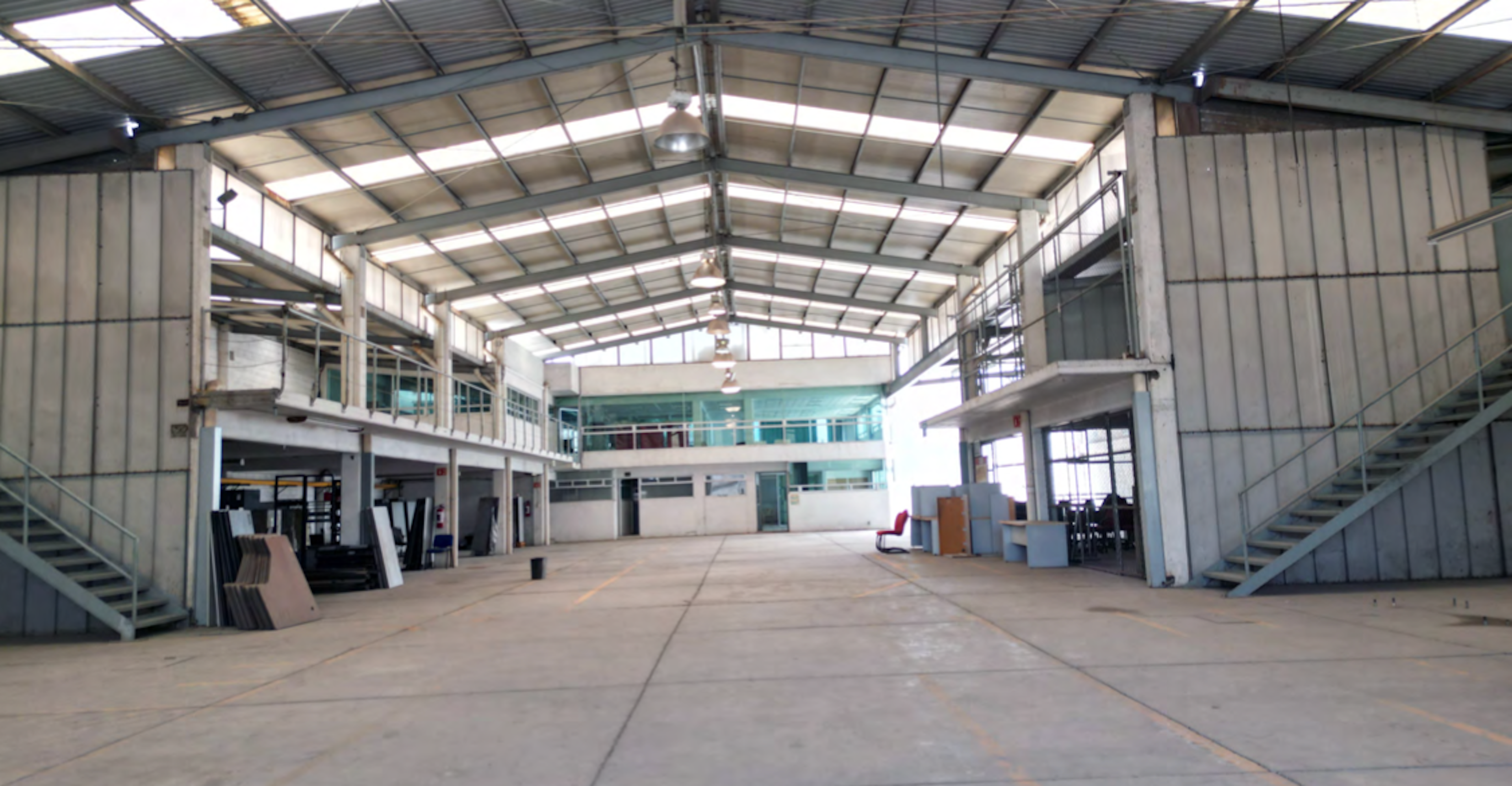La imagen muestra un amplio espacio industrial o de almacenamiento con techo alto de metal y estructura visible. Tiene un segundo nivel parcial con oficinas o áreas de trabajo. El piso es de concreto y hay escaleras metálicas visibles. El espacio parece estar en proceso de remodelación o readaptación, con algunos elementos de construcción y muebles dispersos. La iluminación natural proviene de tragaluces en el techo.