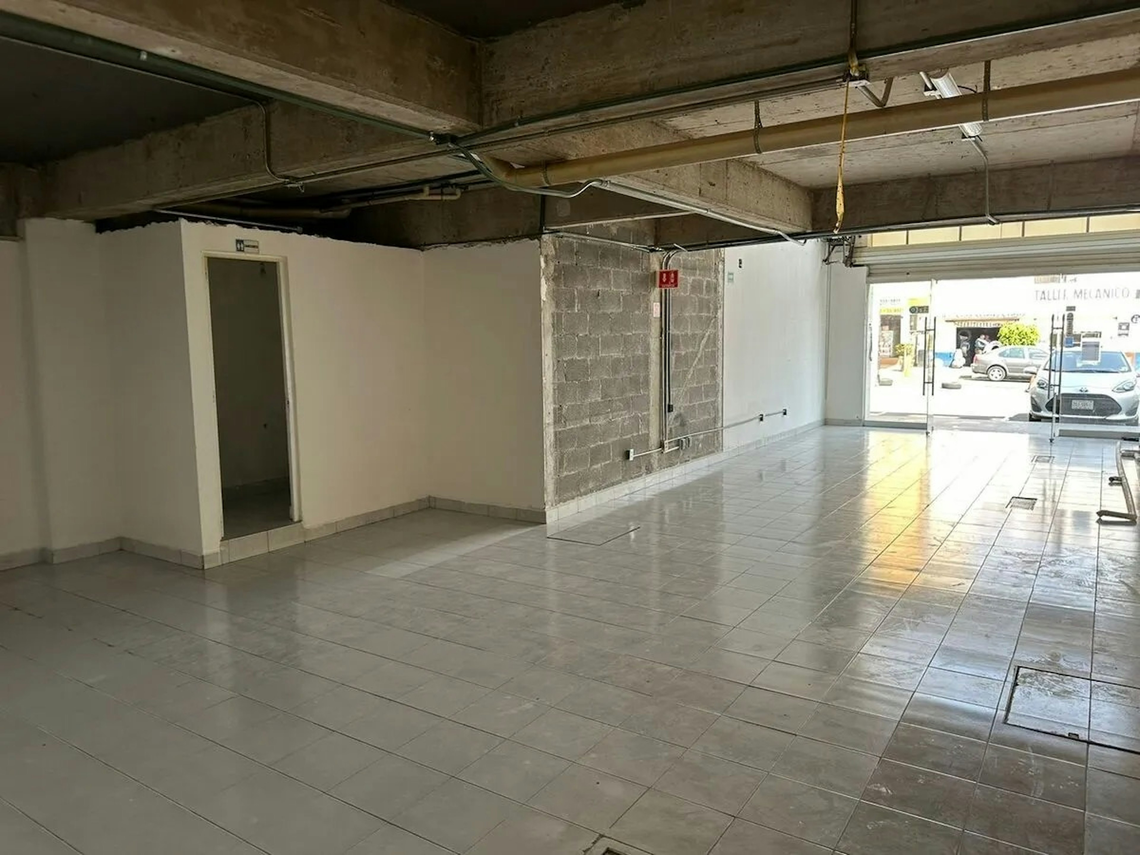 La imagen muestra un local comercial en proceso de remodelación. Se observa un espacio amplio con piso de baldosas, paredes parcialmente terminadas y techo con instalaciones expuestas. Hay una puerta blanca a la izquierda y una entrada de vidrio al fondo que da a la calle. El local parece estar en una etapa avanzada de construcción, listo para ser acondicionado según las necesidades del inquilino.