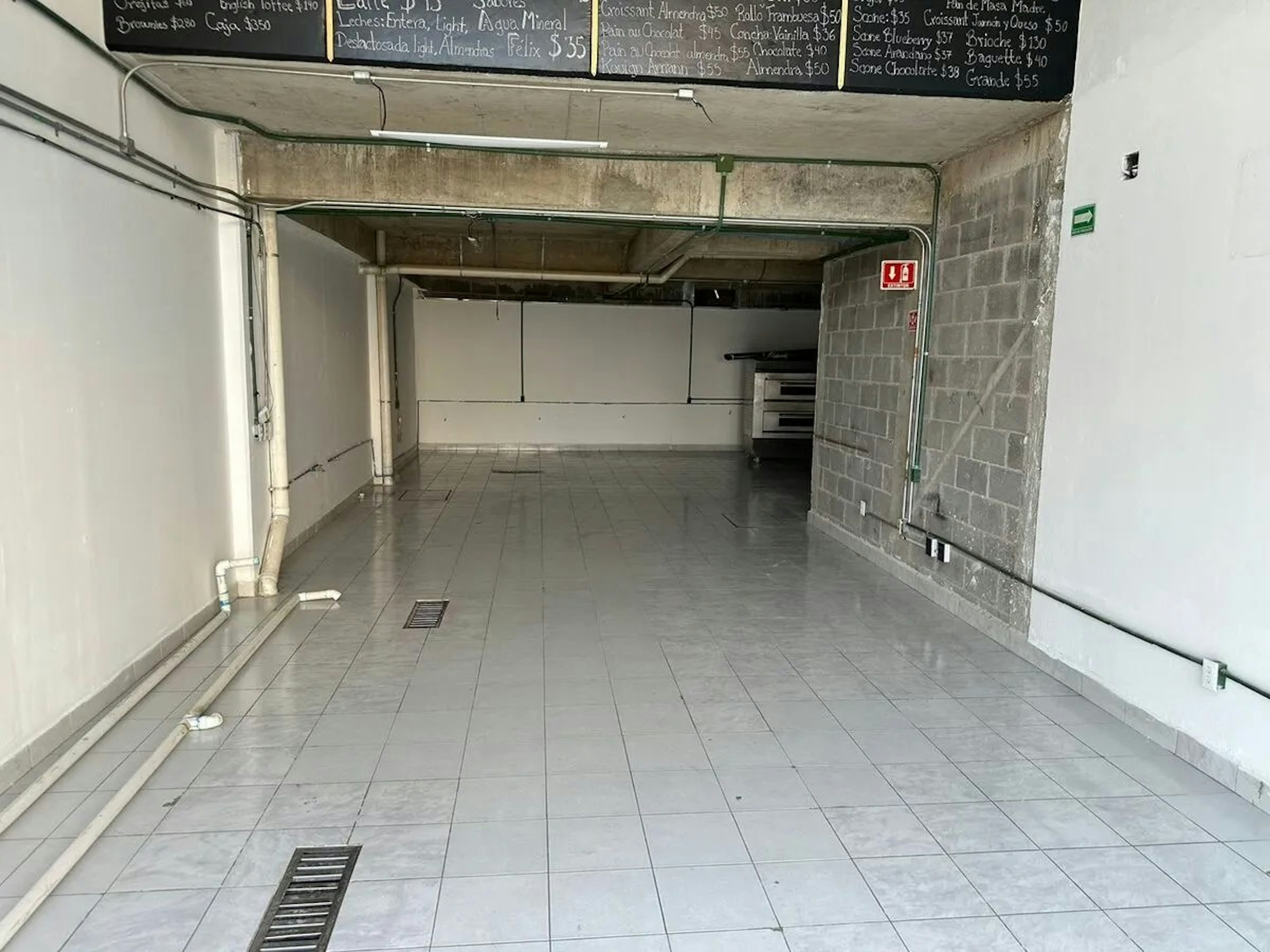 La imagen muestra un local comercial vacío en estado de obra. Tiene un piso de baldosas grises, paredes blancas y de bloques de hormigón expuestos, y tuberías visibles. En la parte superior hay pizarras con menús, sugiriendo que podría ser un café o restaurante en construcción. El espacio es amplio y diáfano, con una zona de cocina al fondo. Parece estar en proceso de renovación o preparación para un nuevo negocio.