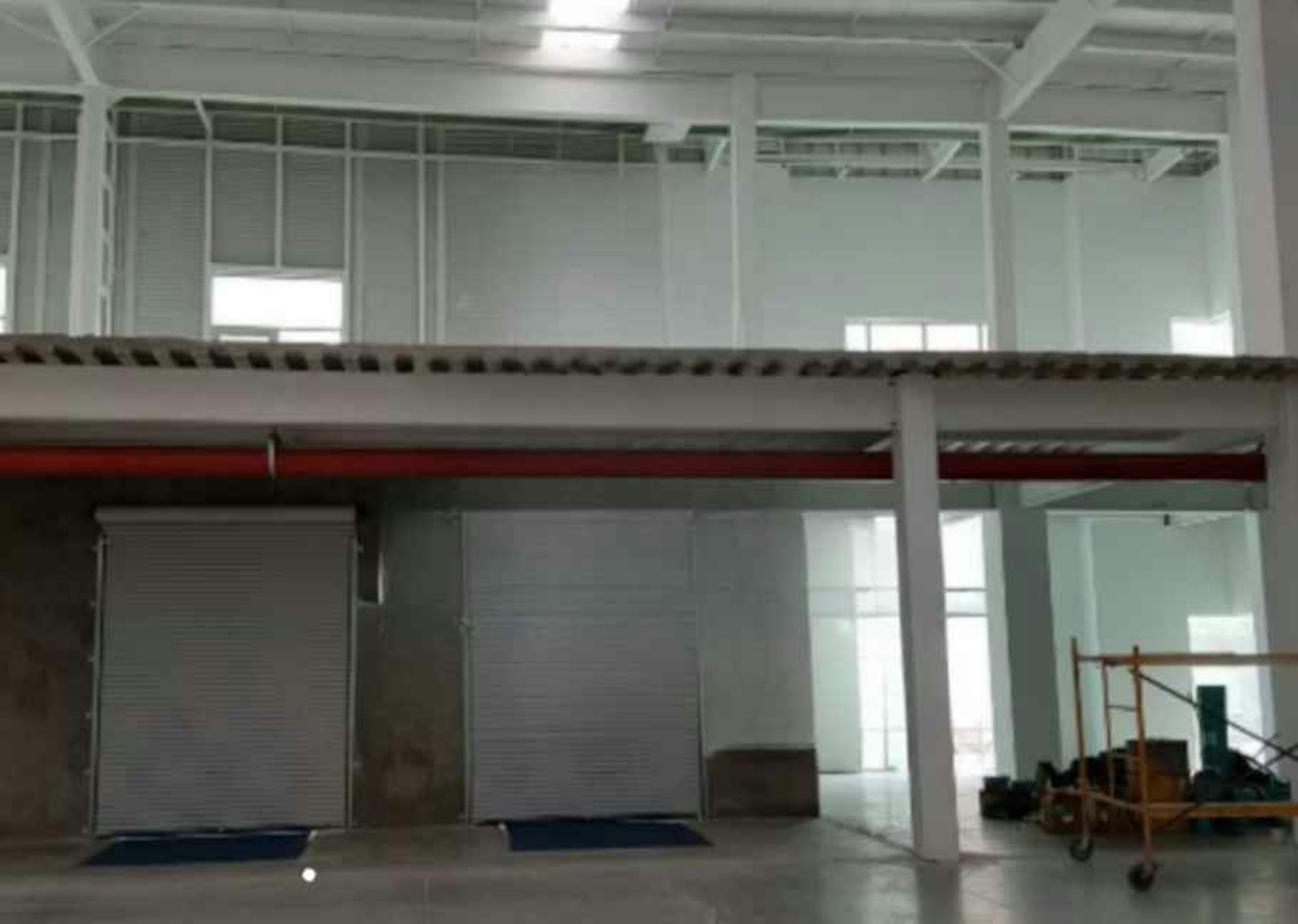 La imagen muestra el interior de un espacio industrial o comercial en construcción. Se aprecia una estructura de dos niveles con techos altos, columnas blancas y vigas expuestas. En la planta baja hay puertas enrollables y áreas sin terminar. El segundo nivel tiene ventanas y barandillas. Se observan algunos materiales de construcción y equipos en el suelo, indicando que el espacio está en proceso de renovación o finalización.