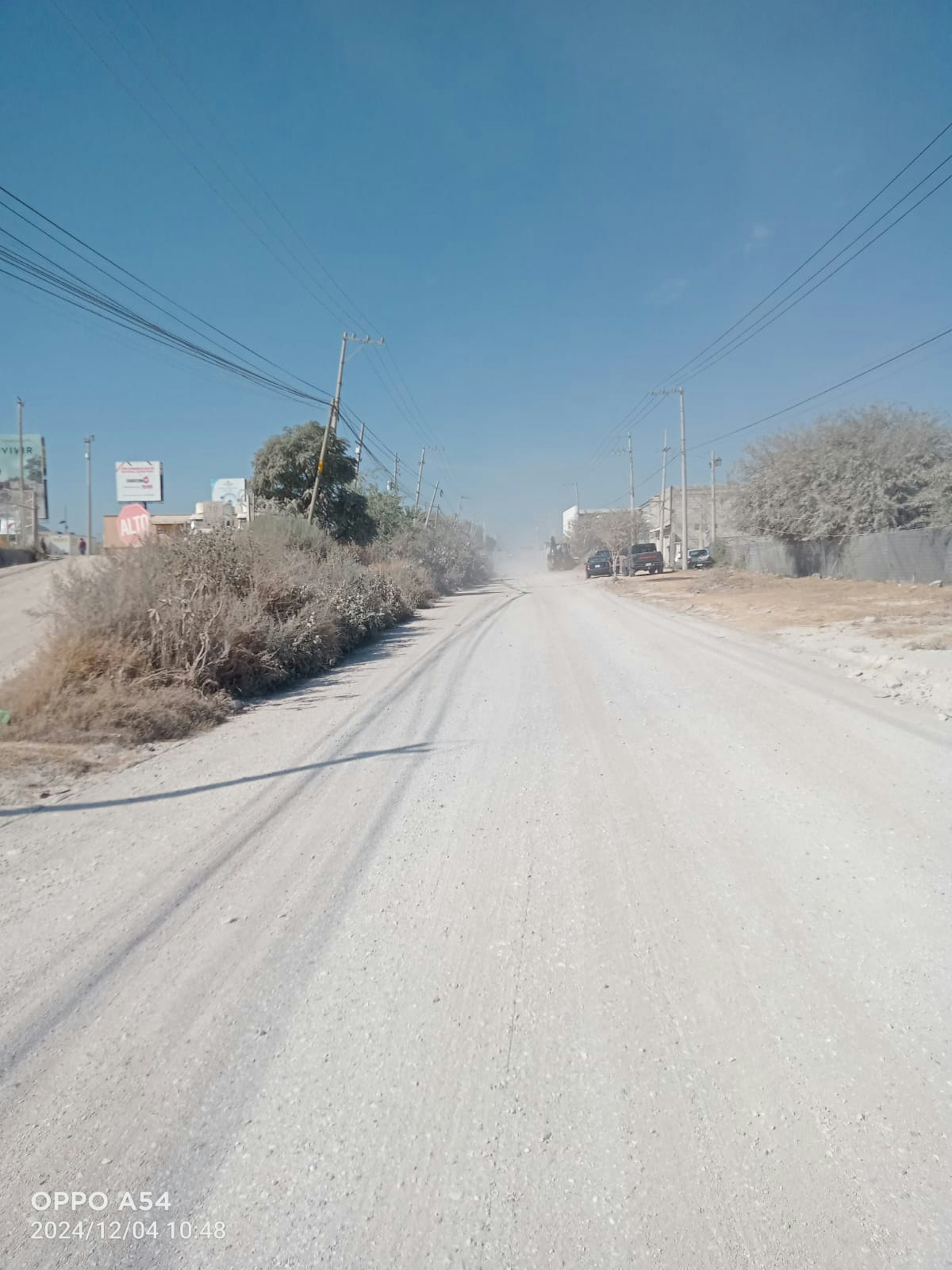 La imagen muestra una calle rural sin pavimentar en un área subdesarrollada. Se observan postes de electricidad, vegetación seca y algunas estructuras básicas a los lados. El camino de tierra sugiere una zona con infraestructura limitada, típica de áreas periféricas o en desarrollo donde podrían surgir oportunidades inmobiliarias para proyectos residenciales o comerciales básicos.