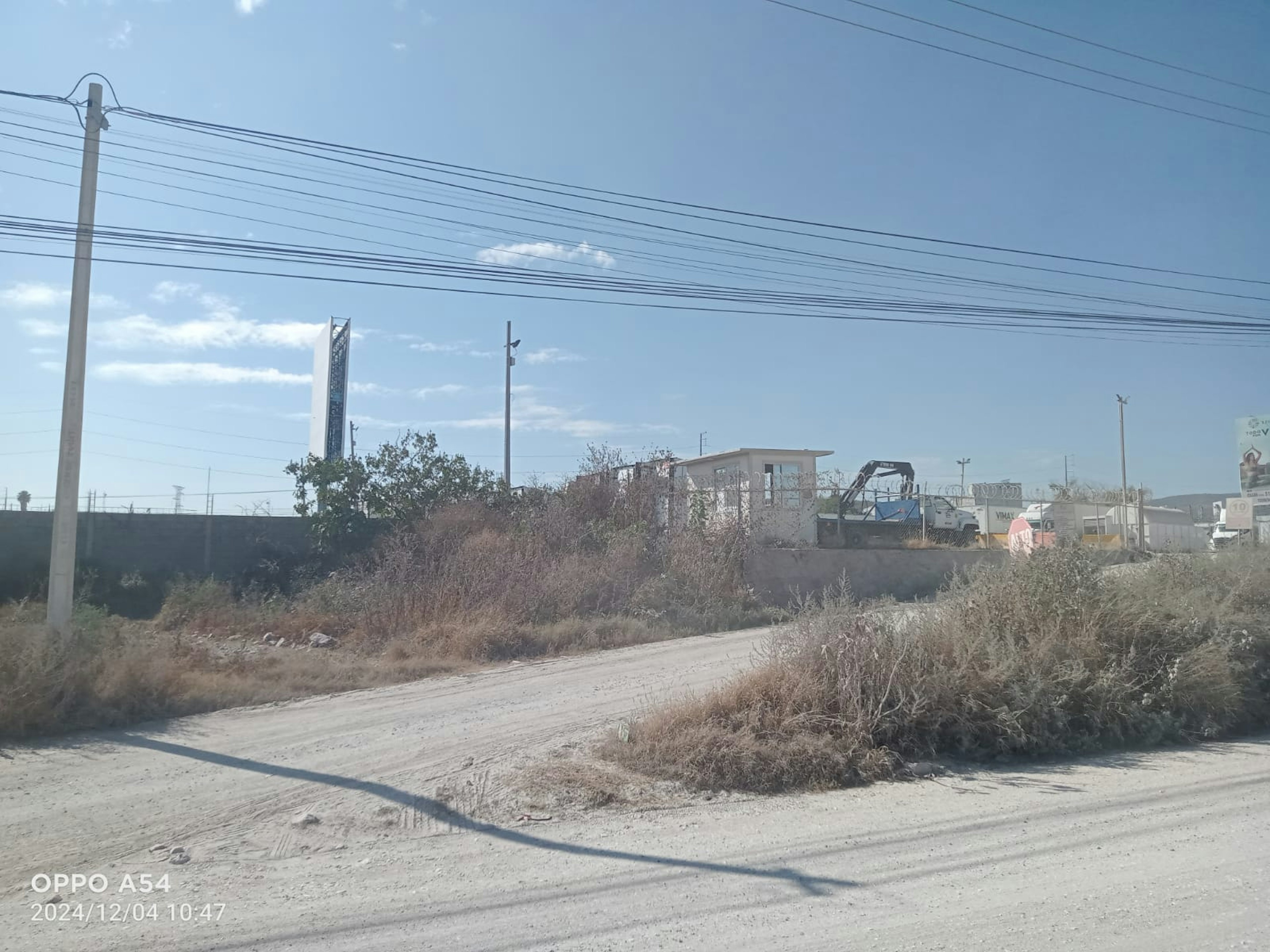 La imagen muestra un terreno baldío en una zona suburbana o industrial. Se observa un camino de tierra, vegetación seca y estructuras distantes. Hay postes de electricidad y cables que cruzan el cielo. Al fondo se ve una torre alta, posiblemente de comunicaciones. El área parece estar en desarrollo, con potencial para futuros proyectos inmobiliarios o industriales. La escena transmite una sensación de espacios abiertos y crecimiento urbano en proceso.