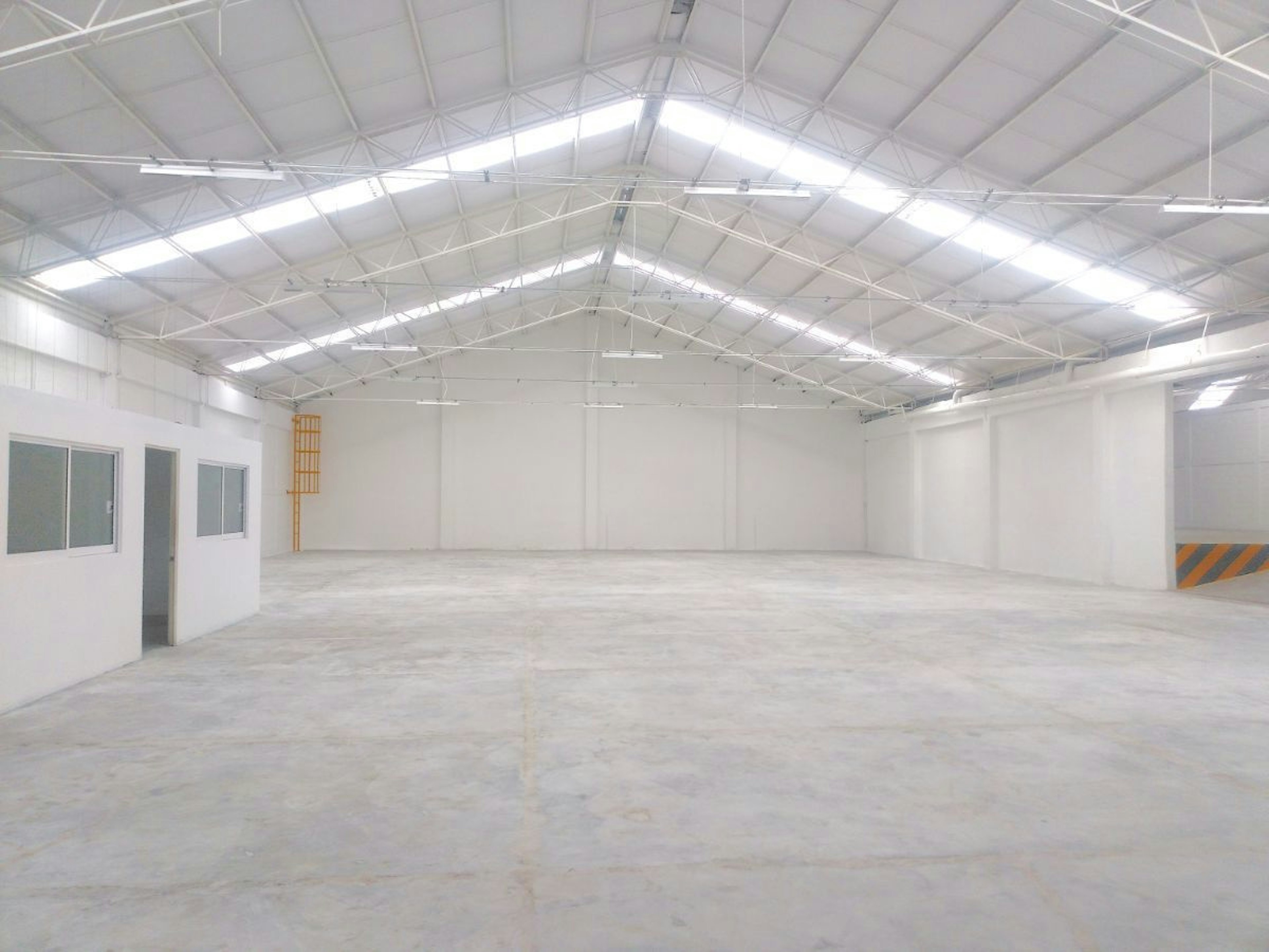 La imagen muestra un amplio espacio industrial o comercial vacío con techo alto de estructura metálica y tragaluces. Las paredes y el suelo son de color claro, probablemente concreto. Se observan algunas ventanas y una puerta en un lado. El espacio está bien iluminado y parece recién construido o renovado, ideal para almacenamiento, manufactura o como área de trabajo multifuncional.