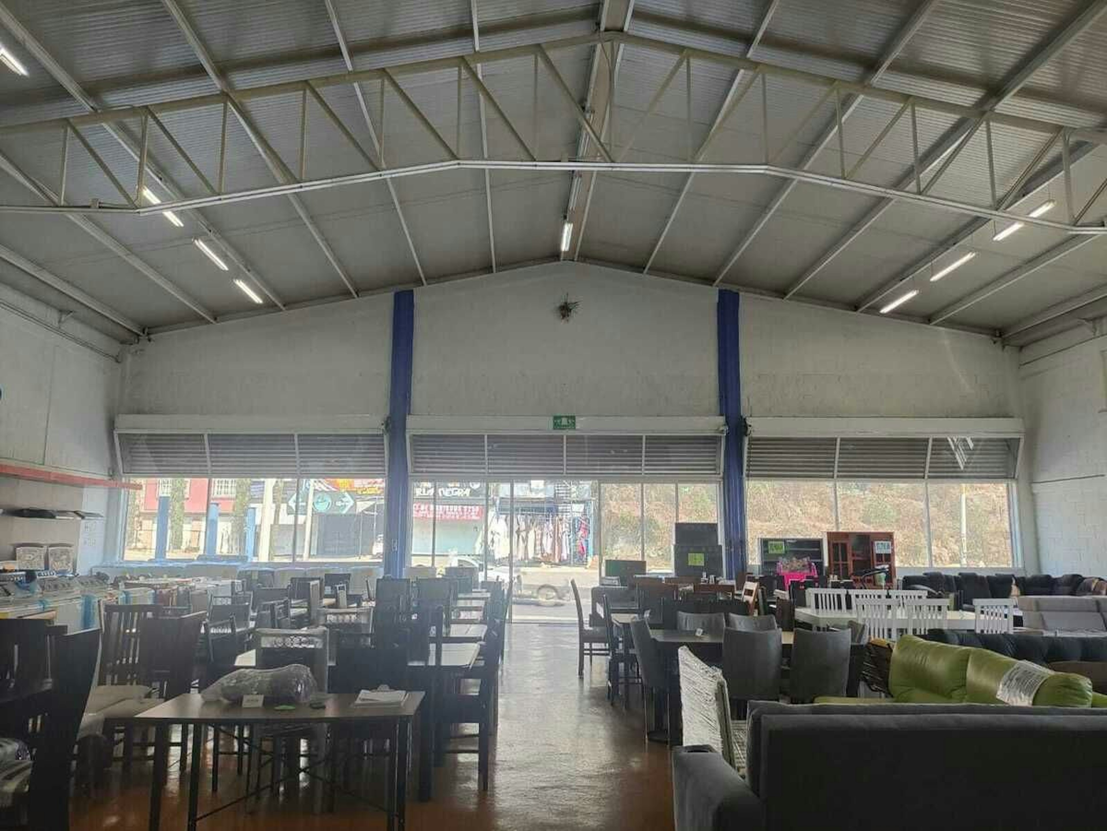 La imagen muestra un amplio espacio interior tipo nave industrial o almacén, convertido en una sala de exposición de muebles. El techo es alto y metálico con iluminación fluorescente. Las paredes son blancas con columnas azules. Hay numerosas mesas, sillas y sofás distribuidos por el suelo, que parece ser de madera. Al fondo se ven grandes ventanales que dan al exterior, donde se aprecian edificios y vegetación.