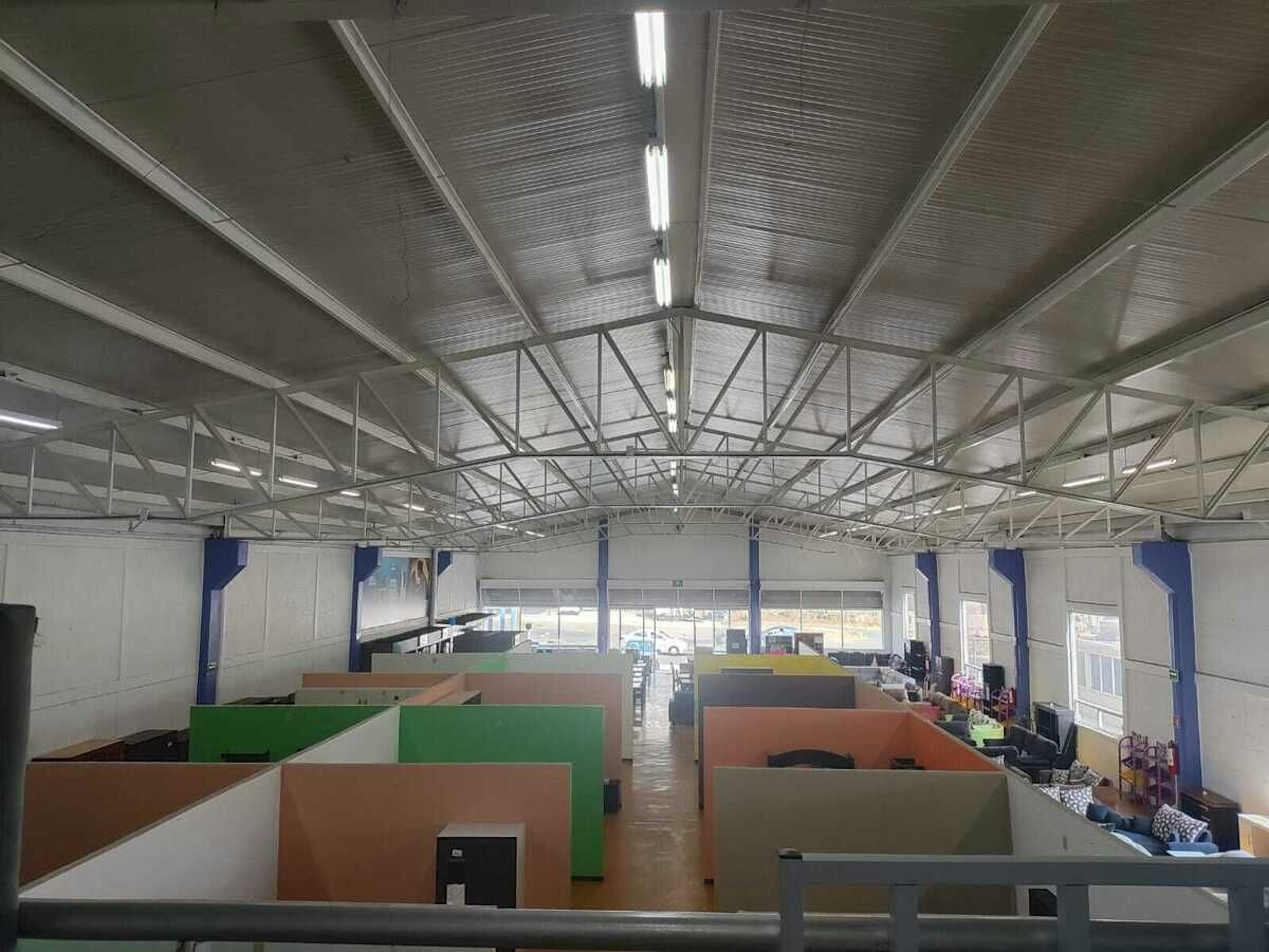 La imagen muestra un amplio espacio de oficina de planta abierta en un edificio industrial. El techo es alto con vigas de metal expuestas y luces fluorescentes. El área está dividida en cubículos de colores brillantes como verde, naranja y gris. Hay ventanas grandes al fondo que permiten la entrada de luz natural. El espacio parece ser un centro de llamadas o una oficina moderna con un diseño flexible y colaborativo.