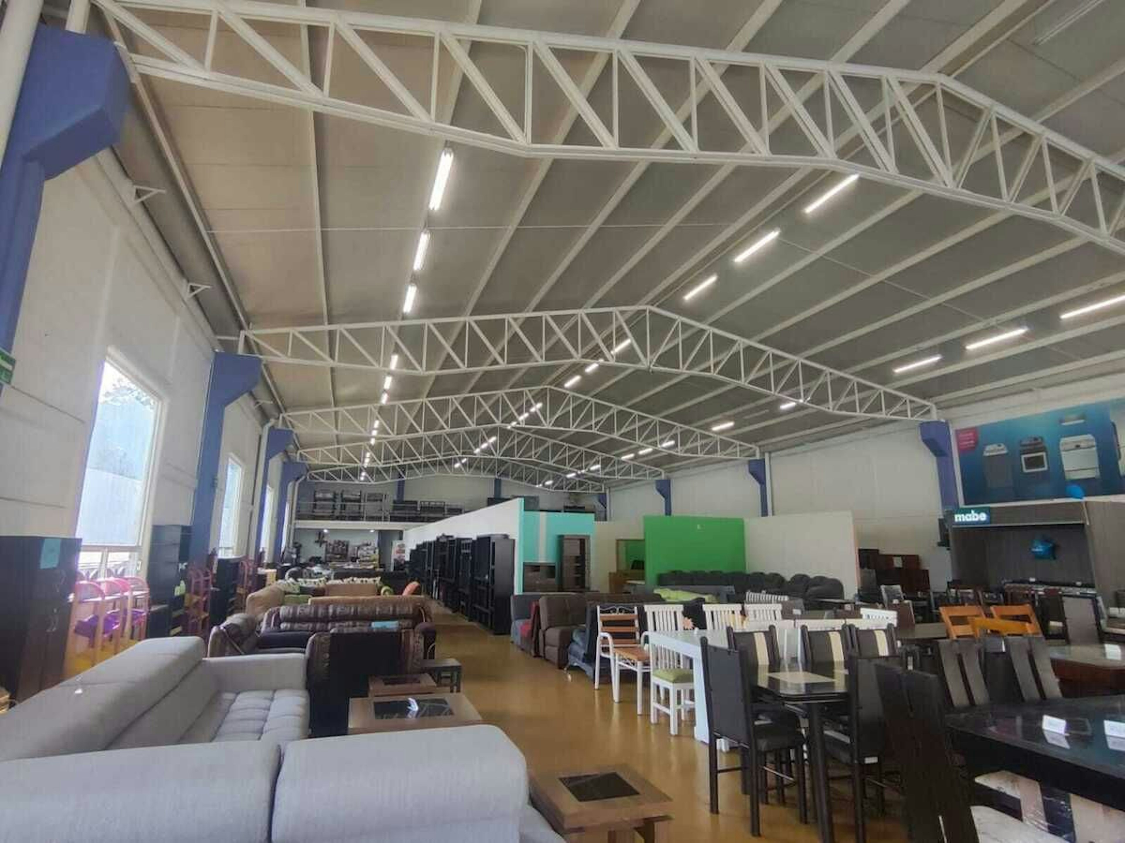 La imagen muestra el interior de un amplio almacén o showroom de muebles y electrodomésticos. El espacio tiene un techo alto con estructura metálica visible y buena iluminación. Se pueden ver diversos tipos de muebles como sofás, sillas y mesas, así como electrodomésticos. El local parece estar bien organizado con secciones para diferentes tipos de productos. El diseño es moderno y espacioso, ideal para exhibir una gran variedad de artículos para el hogar.