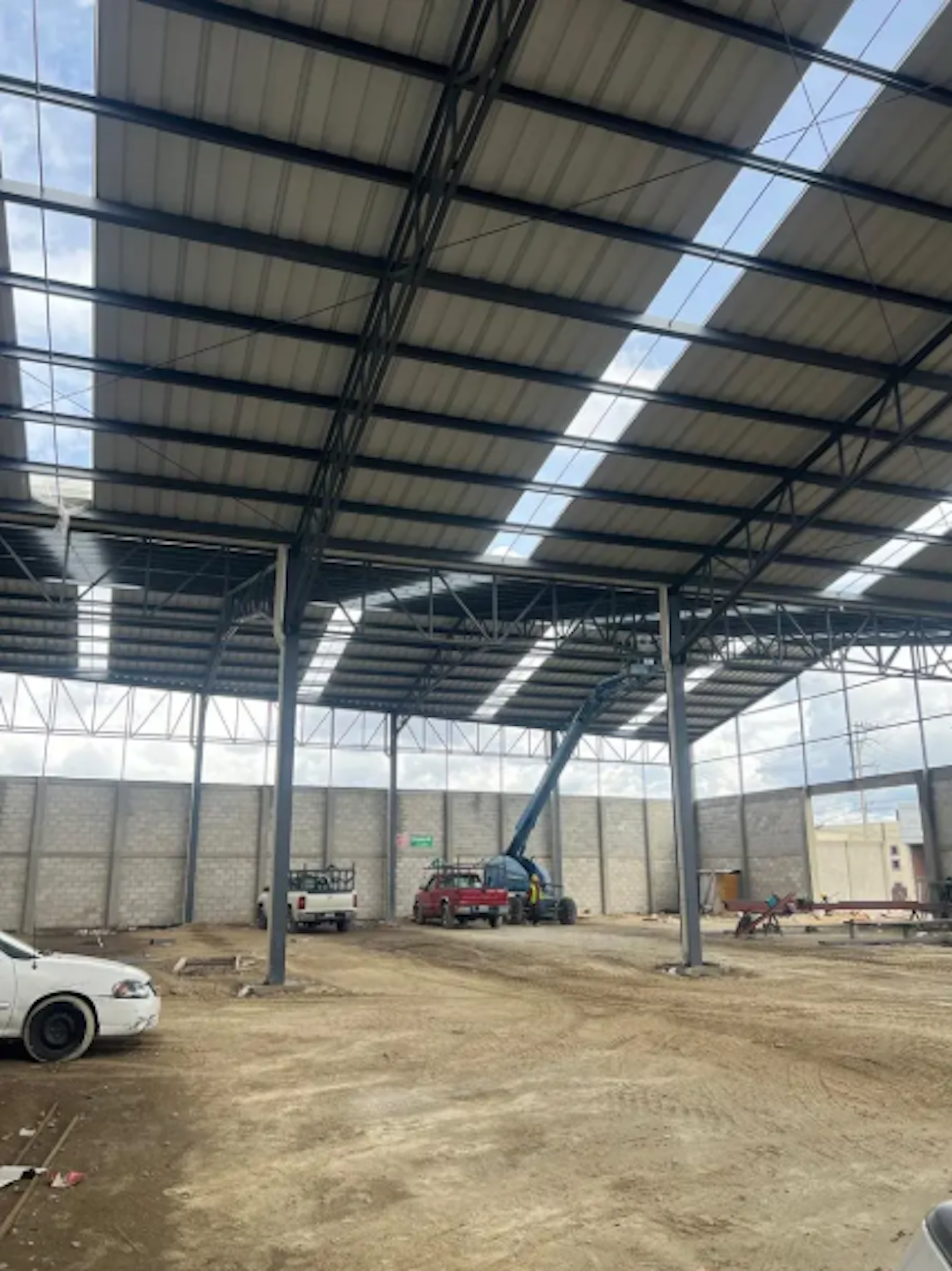 La imagen muestra una estructura industrial en construcción, con un techo de metal alto sostenido por vigas de acero. El espacio interior es amplio y tiene paredes de bloques de concreto. Se observan vehículos estacionados y un elevador hidráulico, indicando que es una obra en progreso. El suelo es de tierra, sugiriendo que el piso aún no está terminado. Este tipo de edificación podría ser utilizada como almacén, fábrica o centro de distribución una vez completada.