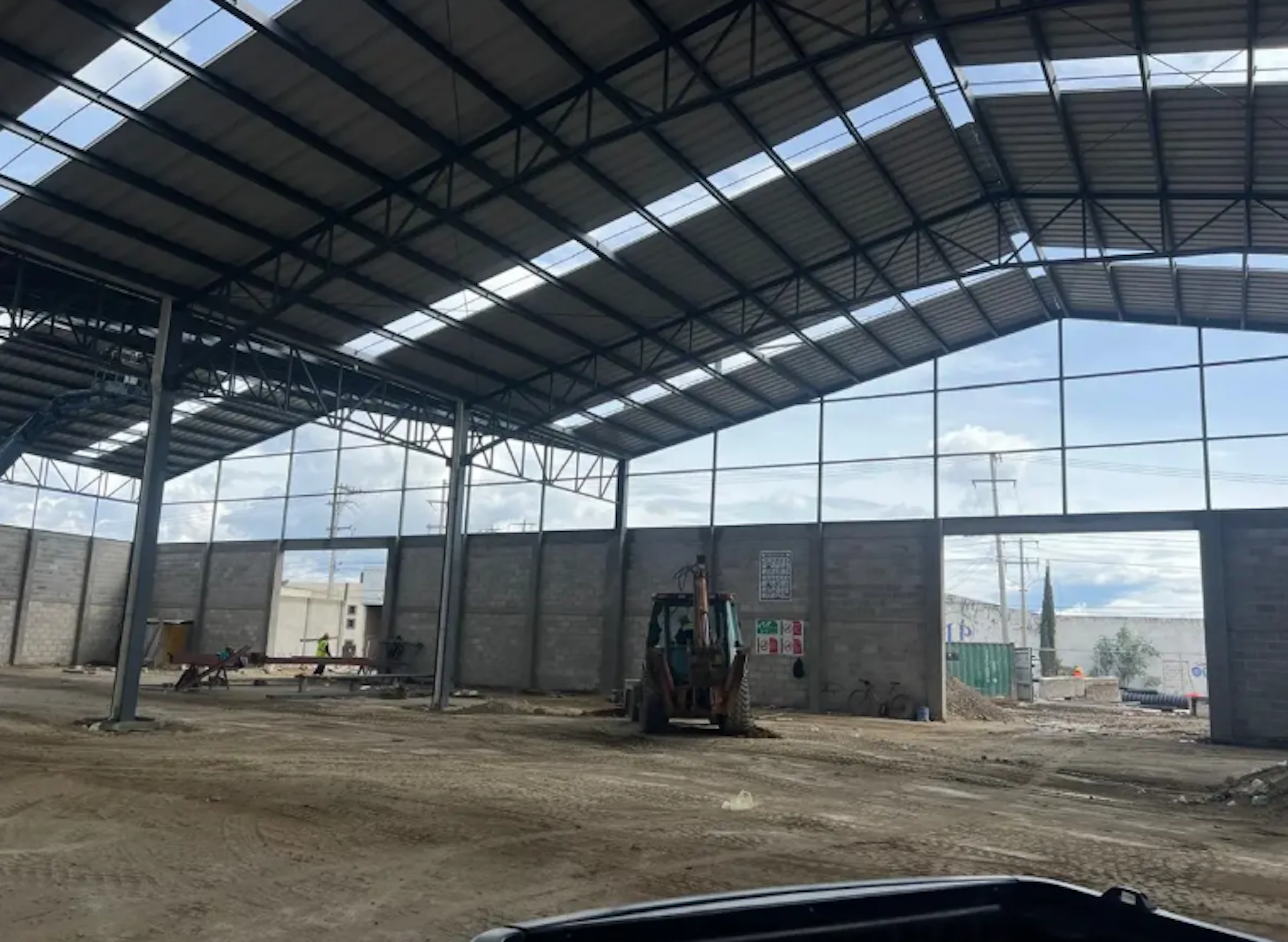 La imagen muestra una estructura industrial en construcción, con un techo de metal arqueado y paredes parcialmente levantadas. El espacio interior es amplio y está en proceso de edificación, con maquinaria pesada visible. El suelo es de tierra y hay señales de trabajo en progreso. La estructura tiene aberturas que permiten ver el exterior, sugiriendo que será un espacio semi-abierto. Este tipo de construcción es común en proyectos de bodegas o naves industriales.