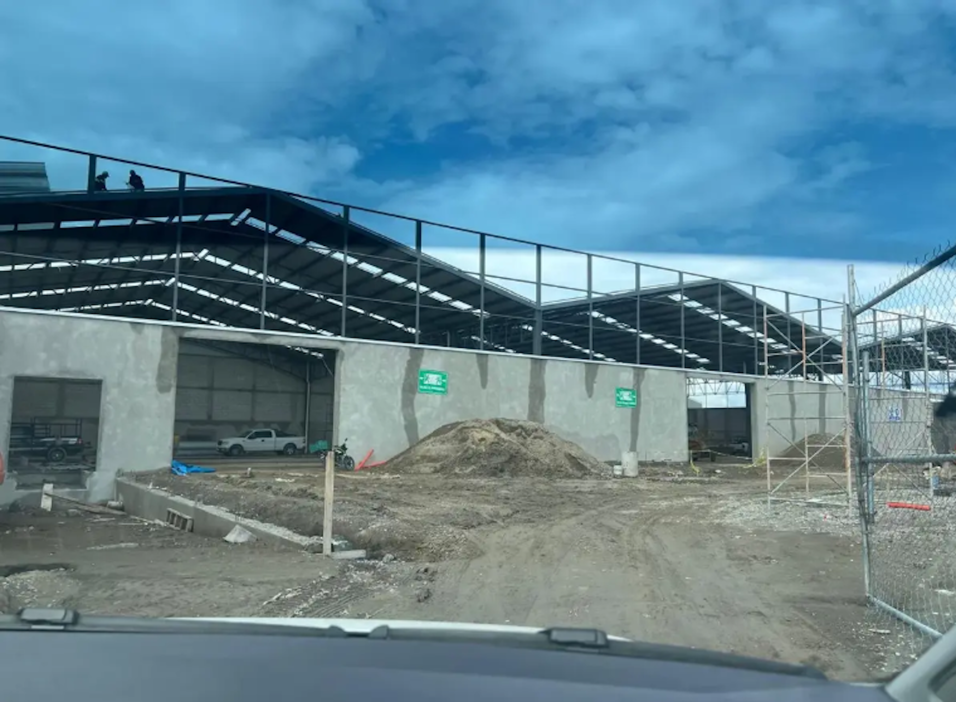 La imagen muestra una construcción en progreso de lo que parece ser un edificio comercial o industrial. Se observa una estructura metálica con techo parcialmente instalado, muros de concreto en la base, y un área de construcción con tierra removida y materiales. El sitio está rodeado por una cerca de seguridad. El cielo es azul con algunas nubes, sugiriendo un día despejado de trabajo en la obra.