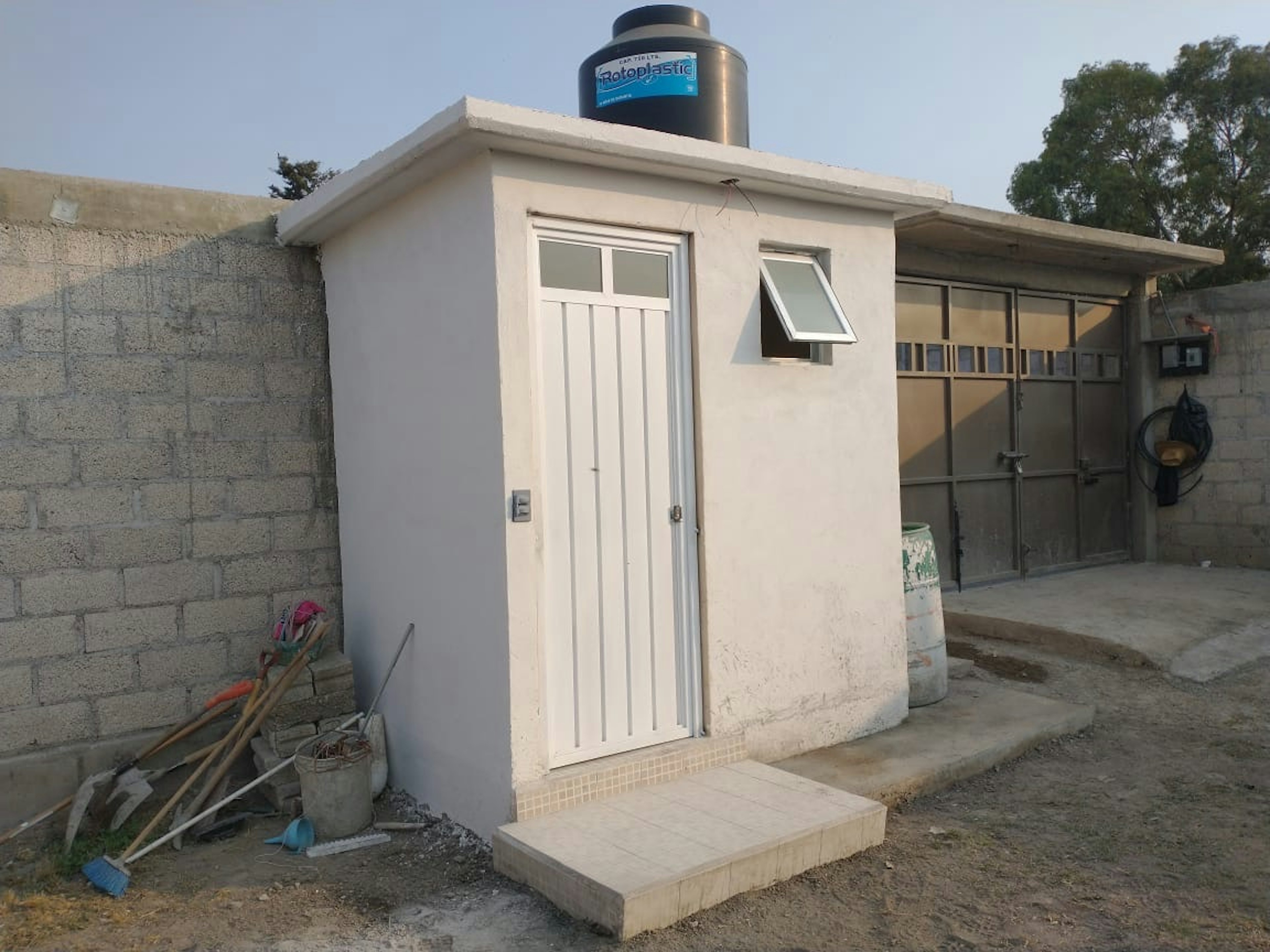 La imagen muestra una pequeña construcción de concreto con techo plano, probablemente un cuarto de servicio o almacén. Tiene una puerta blanca, una ventana pequeña y un tanque de agua Rotoplastic en el techo. Está rodeada por paredes de bloques de concreto y hay herramientas de construcción visibles. El terreno alrededor es de tierra y se ve un área en construcción o remodelación.