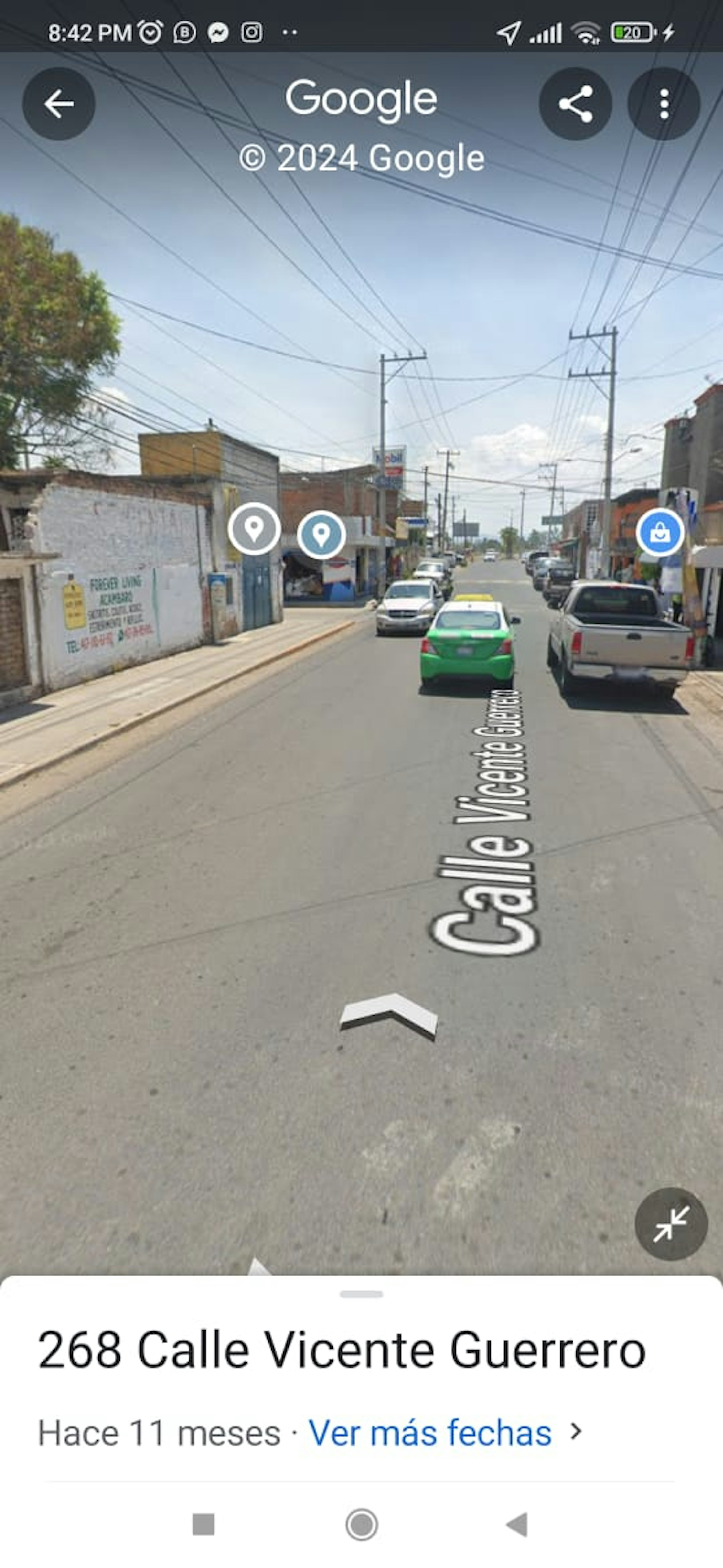 La imagen muestra una calle urbana en una zona aparentemente residencial y comercial mixta. Se ven edificios de baja altura, algunos con negocios en la planta baja. Hay cables eléctricos cruzando la calle y varios vehículos estacionados. La calle está pavimentada y tiene aceras. El entorno sugiere una zona de clase media, posiblemente en una ciudad latinoamericana, con potencial para desarrollo inmobiliario o comercial.