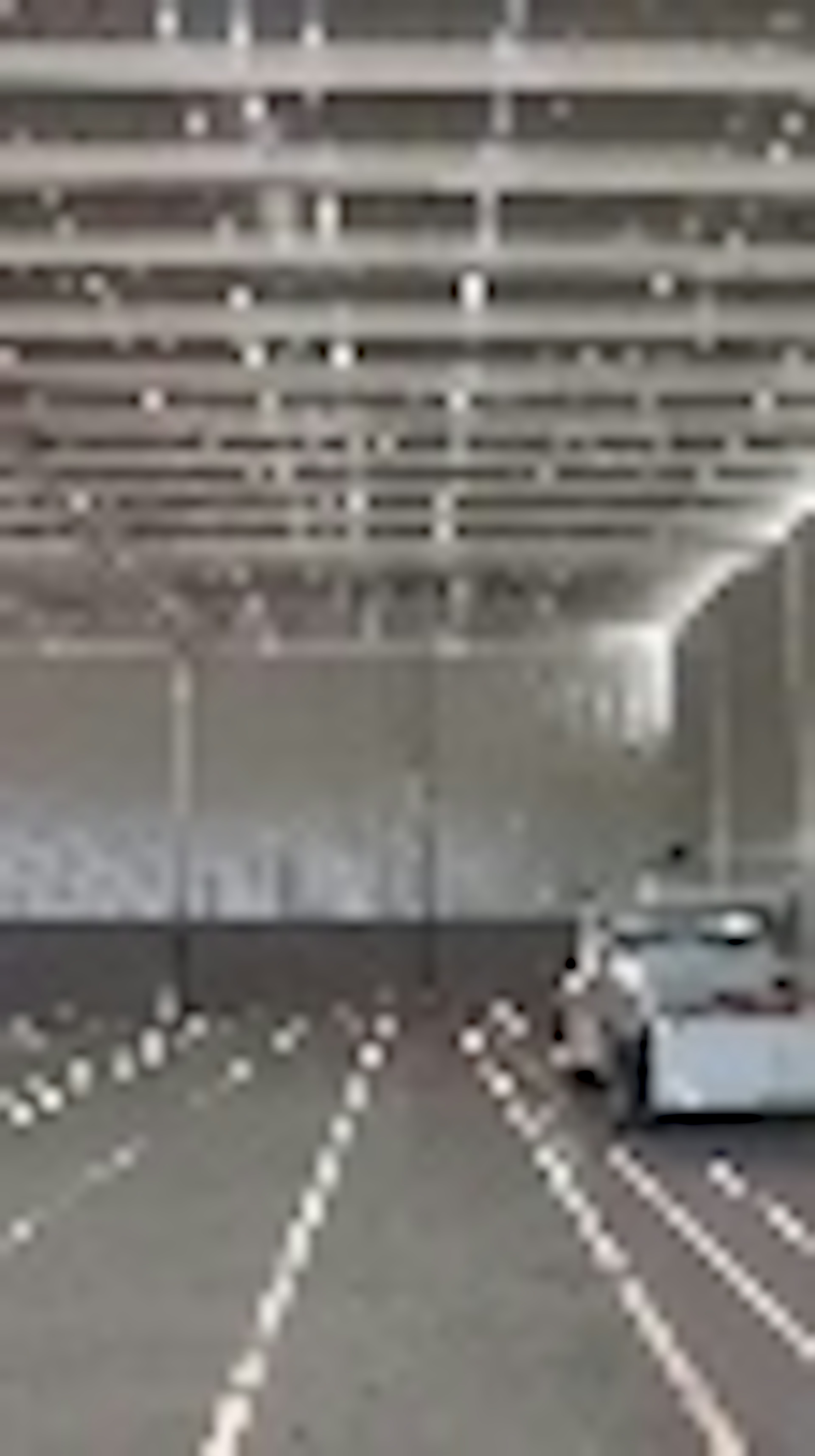 La imagen muestra un gran espacio interior industrial o de almacenamiento. Se ve un techo alto con vigas expuestas y un sistema de iluminación. En el suelo hay líneas blancas pintadas que parecen demarcar áreas o pasillos. En la esquina derecha se aprecia parte de lo que podría ser un vehículo o equipo grande. El espacio parece estar vacío y listo para ser utilizado como almacén o para operaciones logísticas.