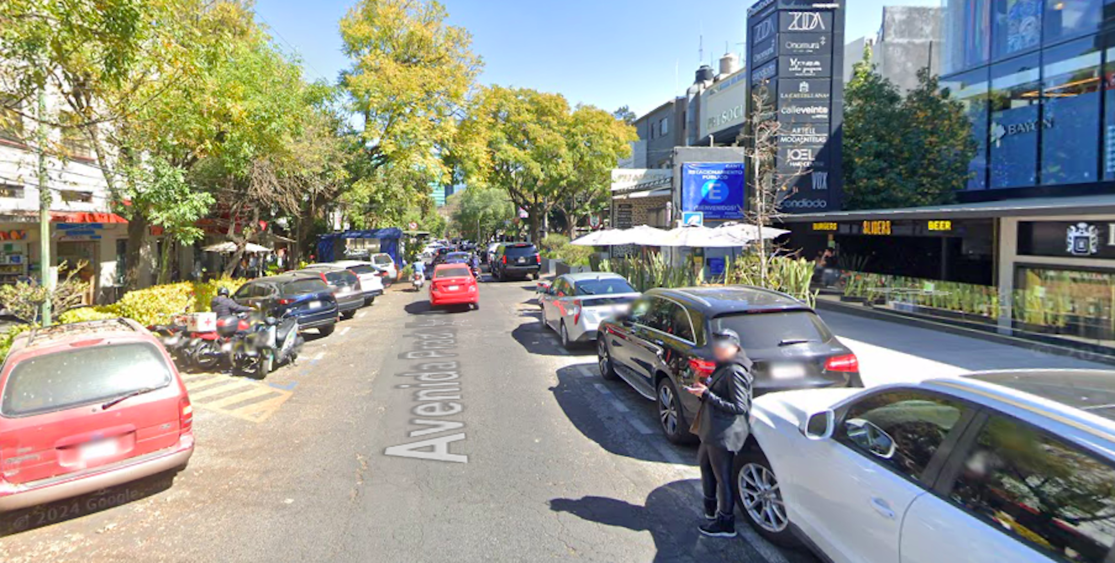 La imagen muestra una calle comercial urbana con edificios de varios pisos, árboles y una variedad de negocios. La avenida está llena de vehículos estacionados a ambos lados y algunos en movimiento. Se observan carteles publicitarios y letreros de negocios. El ambiente sugiere una zona comercial activa y bien establecida, típica de un área urbana de clase media a alta con potencial inmobiliario para locales comerciales y oficinas.