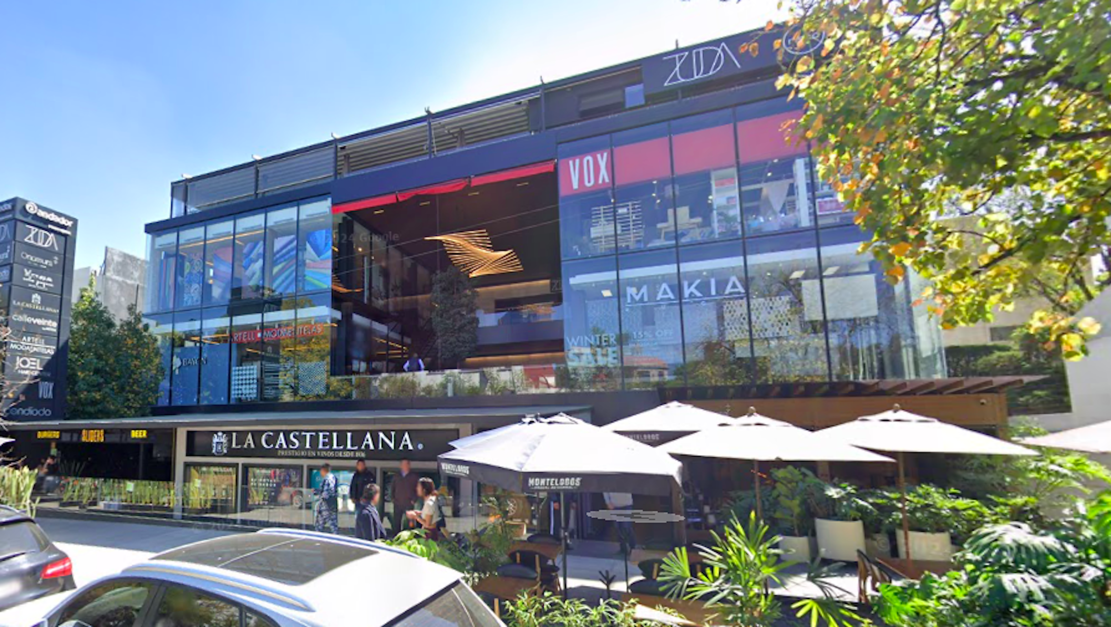 La imagen muestra un moderno centro comercial o complejo de tiendas llamado La Castellana. El edificio tiene una fachada de vidrio con múltiples niveles, donde se pueden ver logotipos de tiendas como VOX y MAKIA. En la planta baja hay restaurantes con terrazas y sombrillas blancas. El diseño es contemporáneo, con una mezcla de materiales como cristal y metal. El entorno incluye vegetación y autos estacionados, sugiriendo una ubicación urbana comercial de alto nivel.