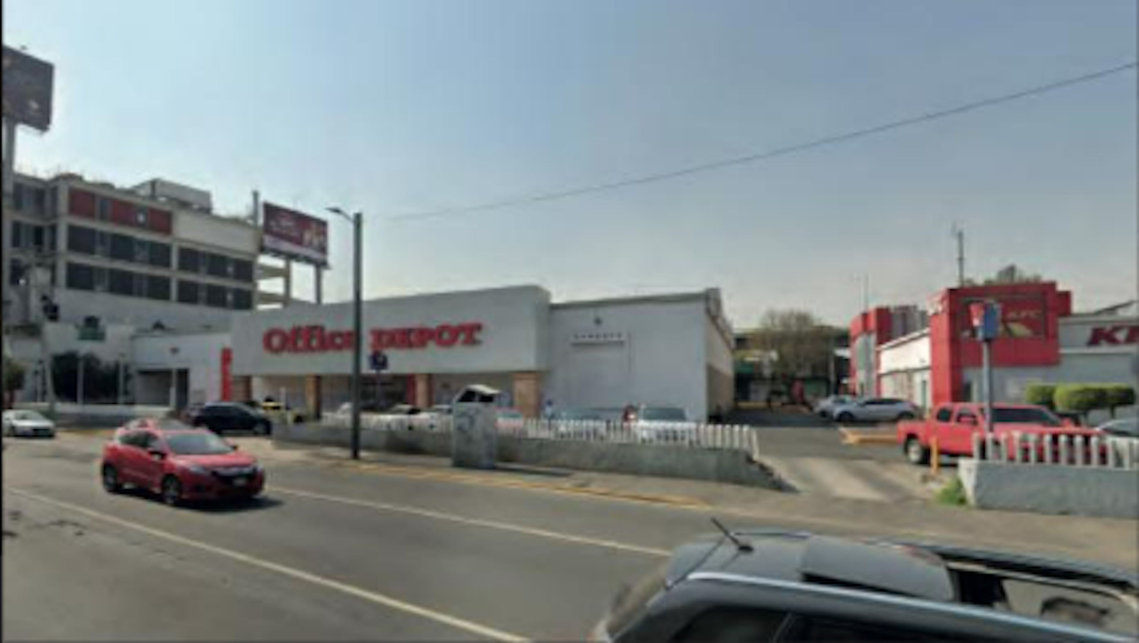 La imagen muestra una calle comercial con un local prominente de Office Depot. Se observan varios edificios comerciales, estacionamientos y algunos vehículos circulando. El área parece ser una zona suburbana o de las afueras de una ciudad, con un cielo despejado. La escena sugiere un área de uso mixto con oficinas y tiendas minoristas.